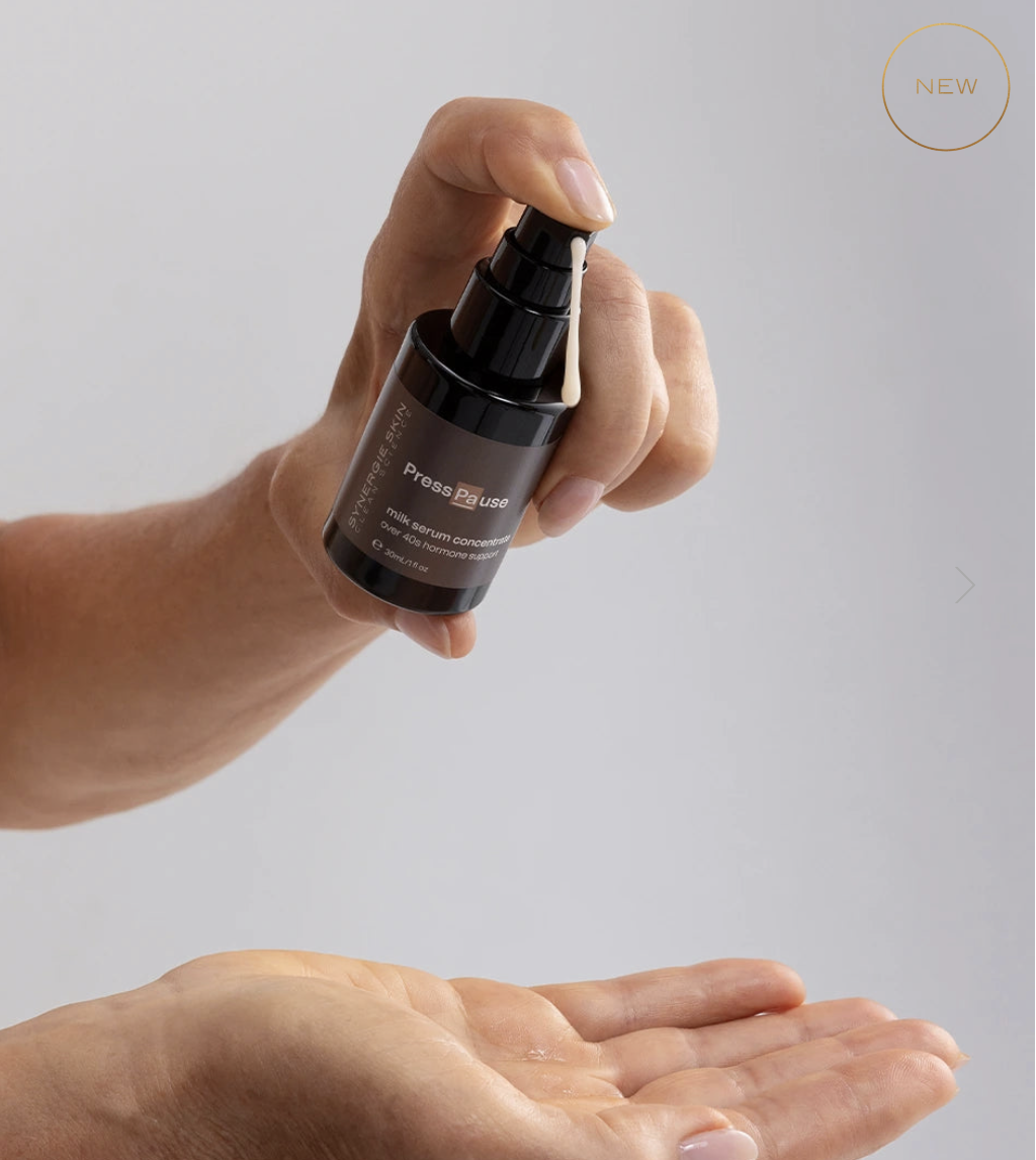 A hand holding a small black bottle labeled "Press Pause" milk serum concentrate with a drop of the serum coming out of the pump dispenser. Another hand is below, ready to receive the serum.