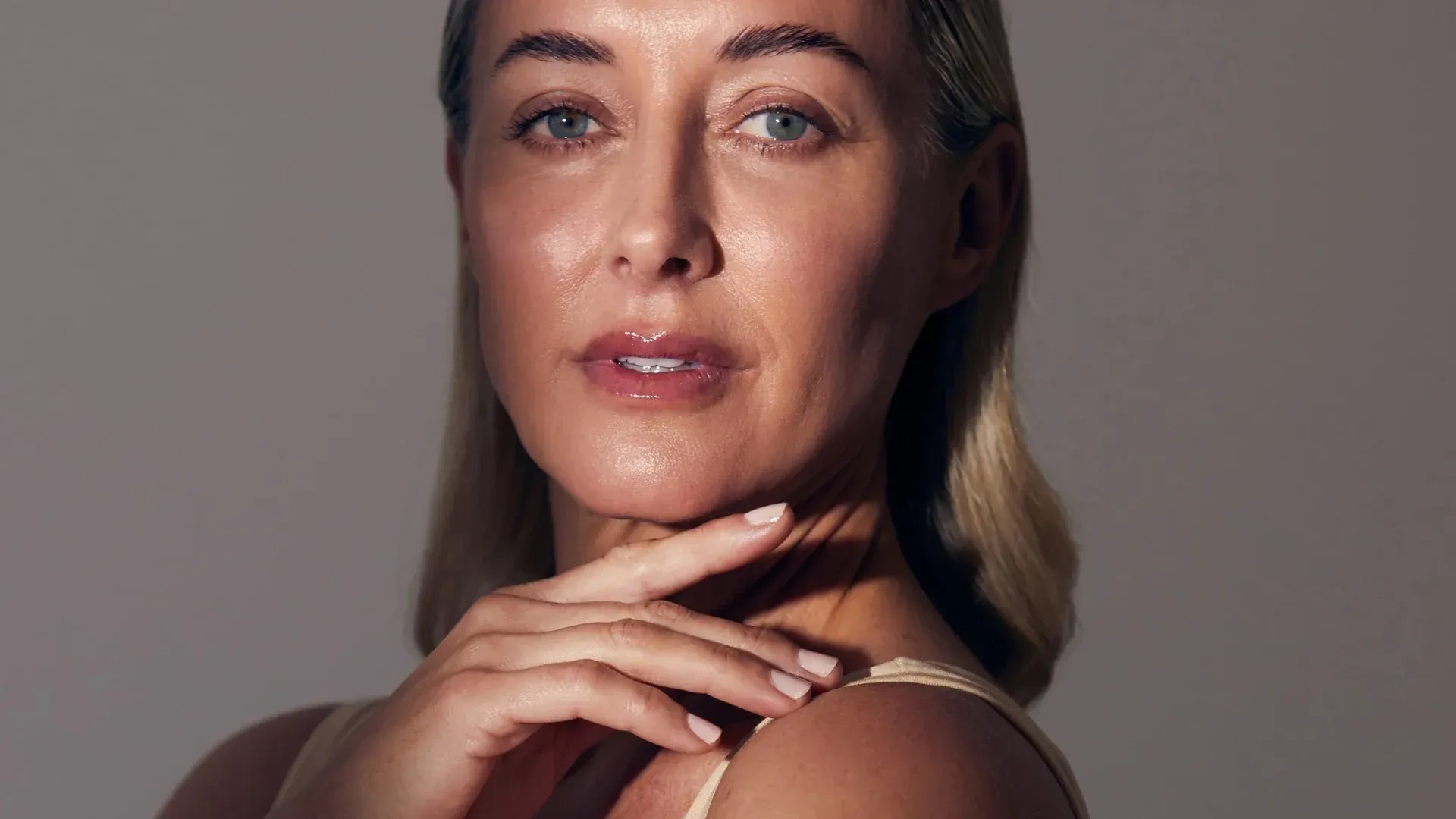 Close-up of a woman with blonde hair and blue eyes, gently touching her neck with her left hand, against a neutral background.