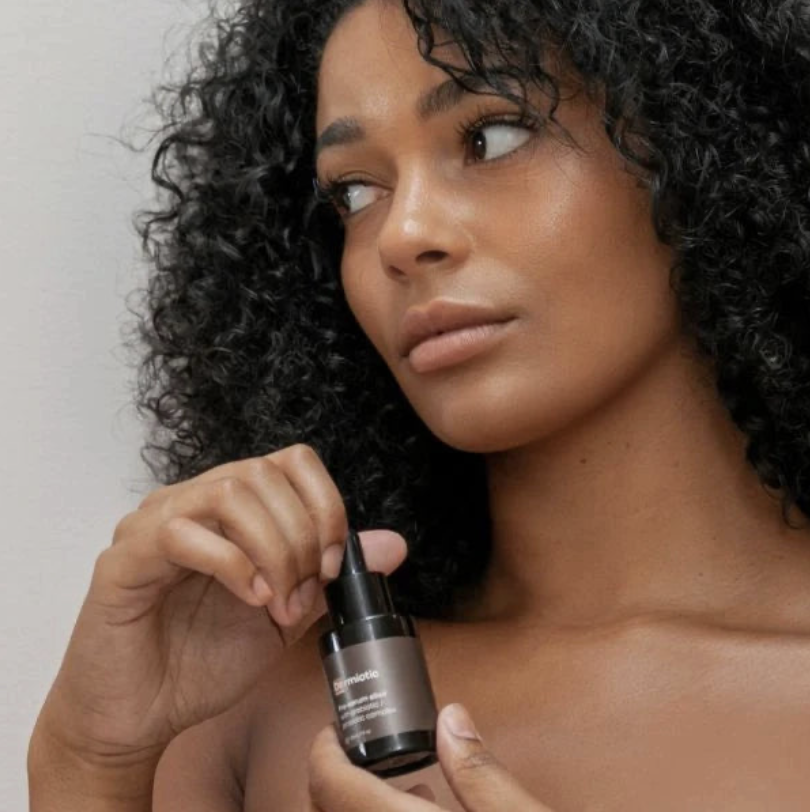 A woman with curly black hair holding a skincare or cosmetic product close to her shoulder, looking to the side.