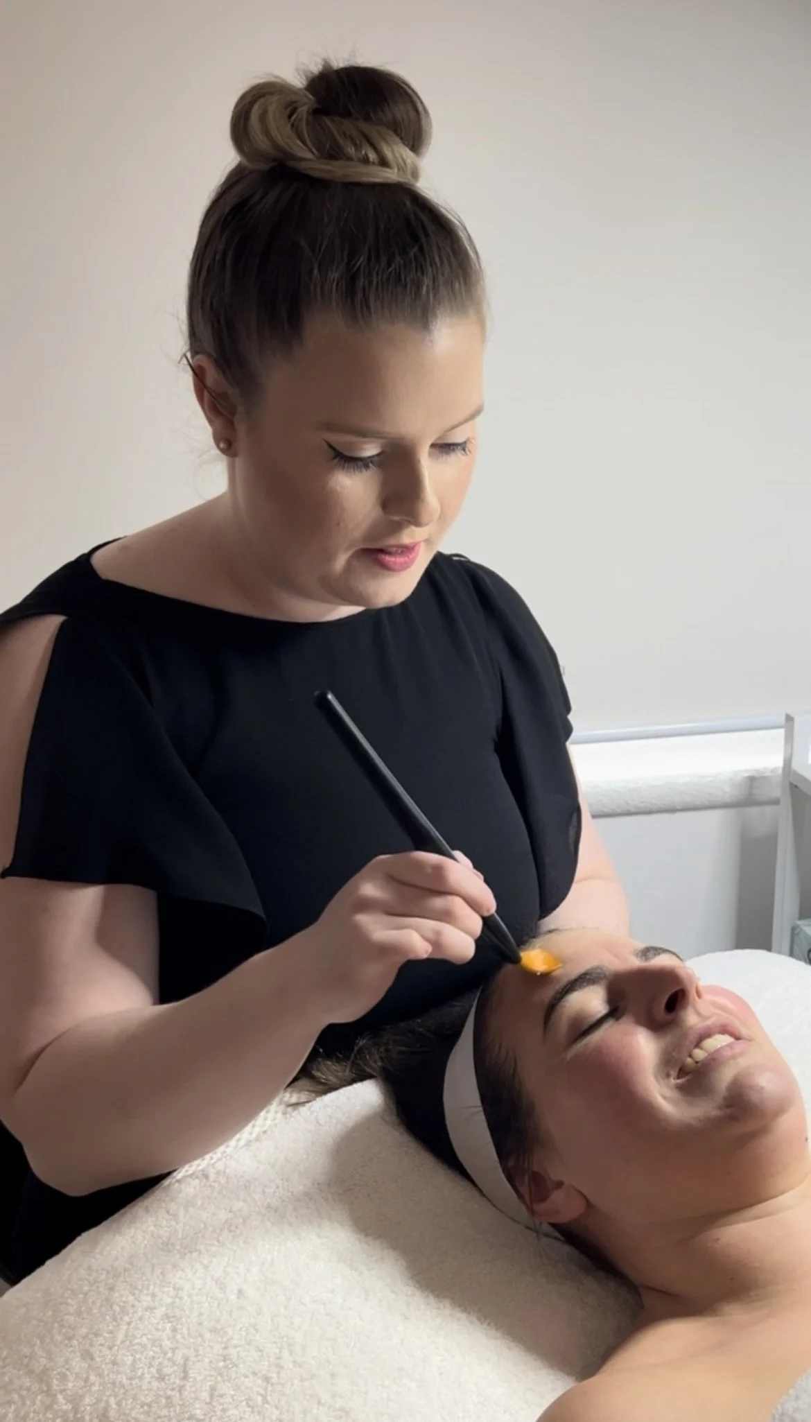 A woman receiving a facial treatment with a tool on her forehead, lying on a treatment bed, smiling with her eyes closed.