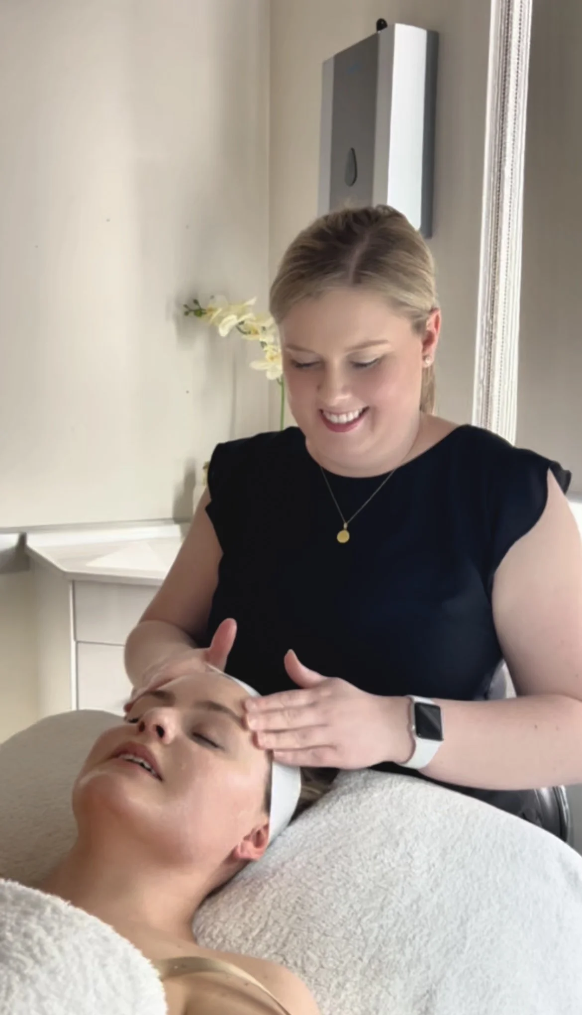 A woman giving a facial massage to a woman lying on a treatment bed during a spa or skincare session.
