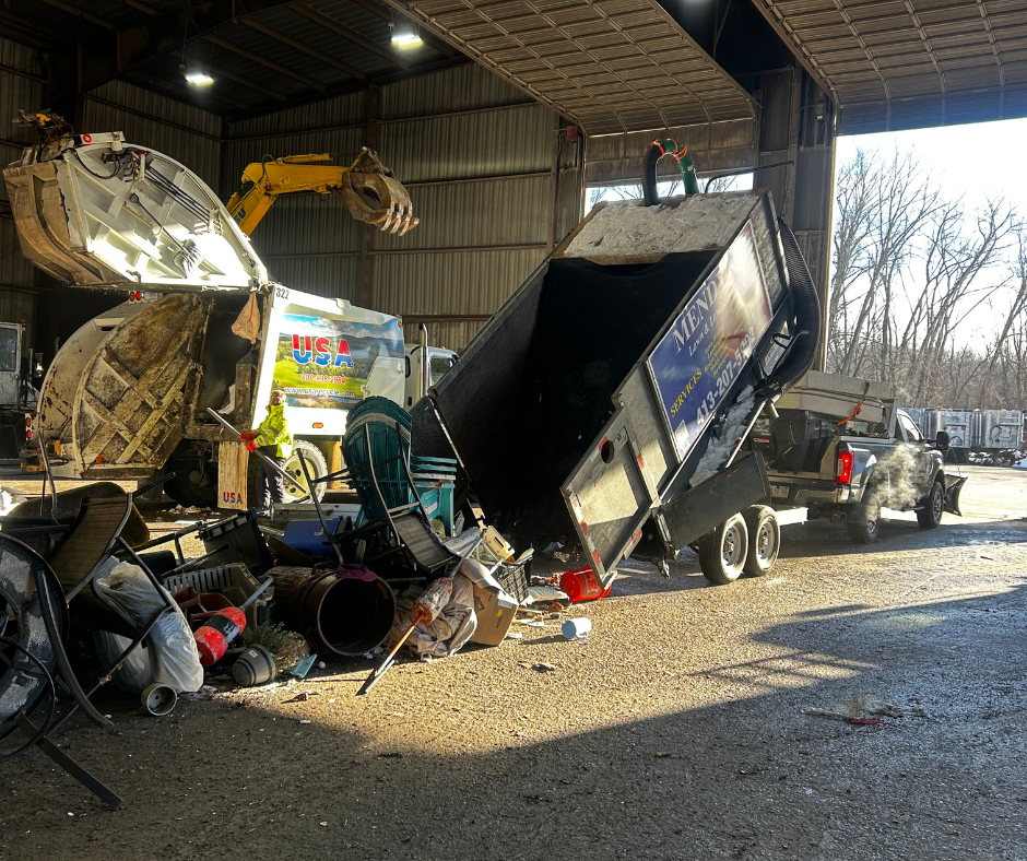 A large vehicle trailer is tilted with its contents spilling onto the ground inside a warehouse or garage. There are miscellaneous items including garbage bins, tools, and debris scattered on the ground. A person in a yellow safety vest is standing near a large vacuum truck, and other trucks and equipment are visible outside and inside the structure.