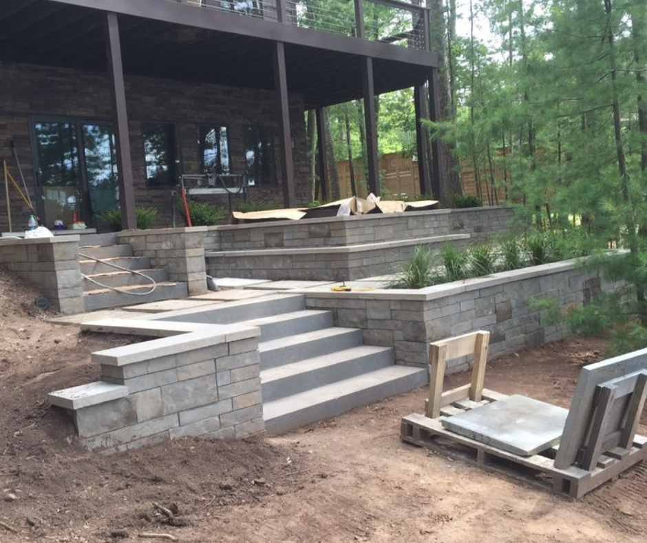 Under construction outdoor patio with stone stairs and retaining walls, surrounded by trees.