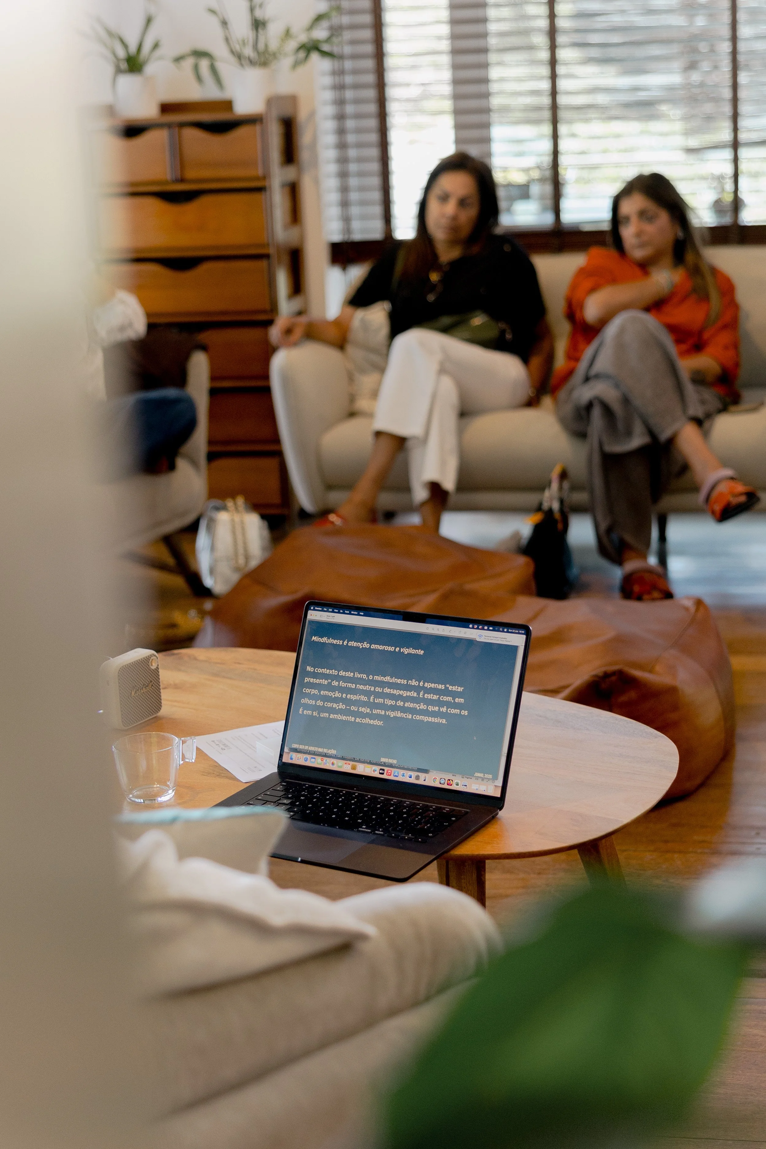A laptop on a wooden table displaying a text about mindfulness, with a small speaker, a glass, and some papers nearby. In the background, two women are sitting on a white sofa, engaged in conversation in a cozy, well-lit living room with wooden floors and window blinds.
