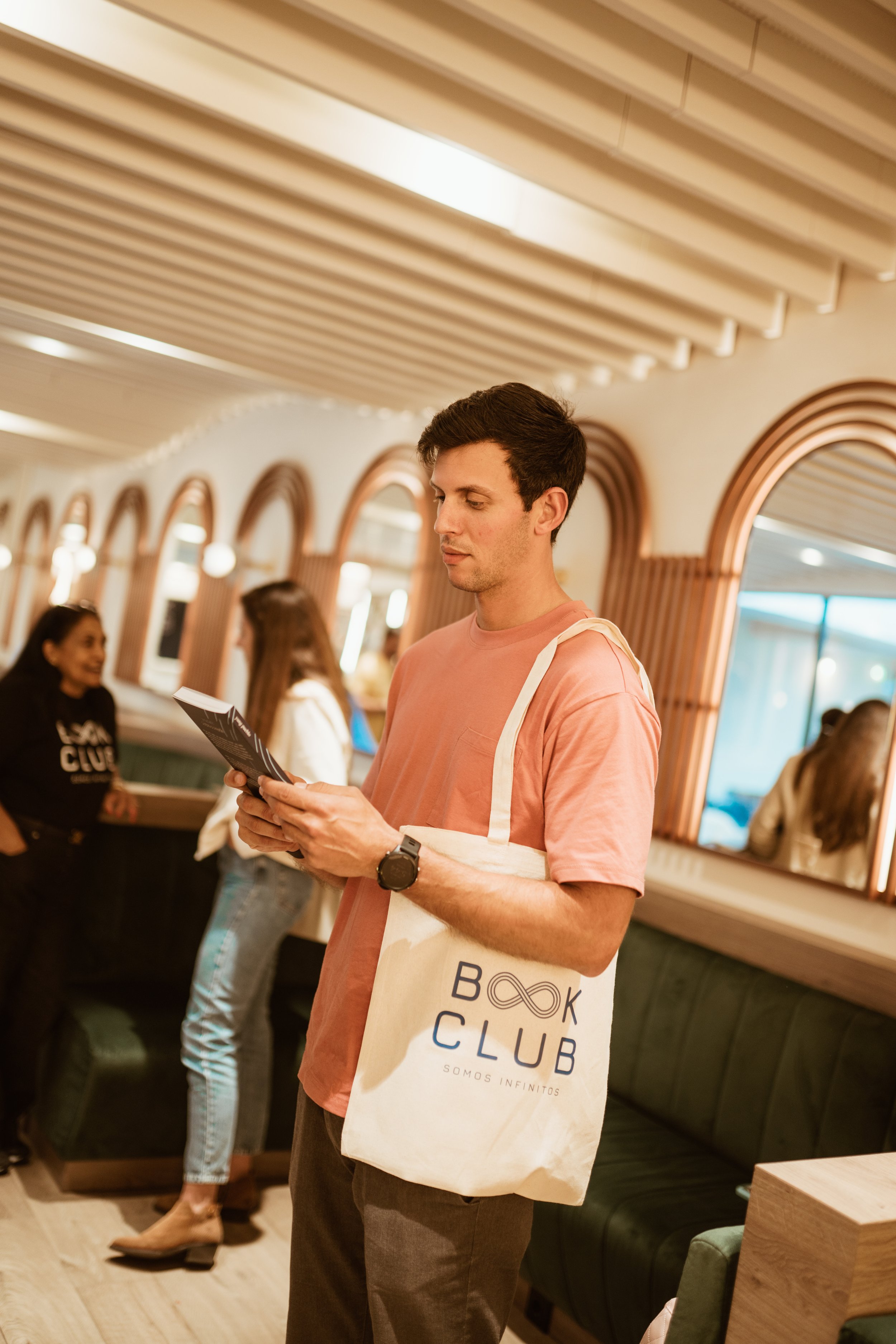 A young man wearing a pink t-shirt and wristwatch, holding a book and a tote bag with "Book Club" written on it, in a lively cafe or restaurant with other people in the background.