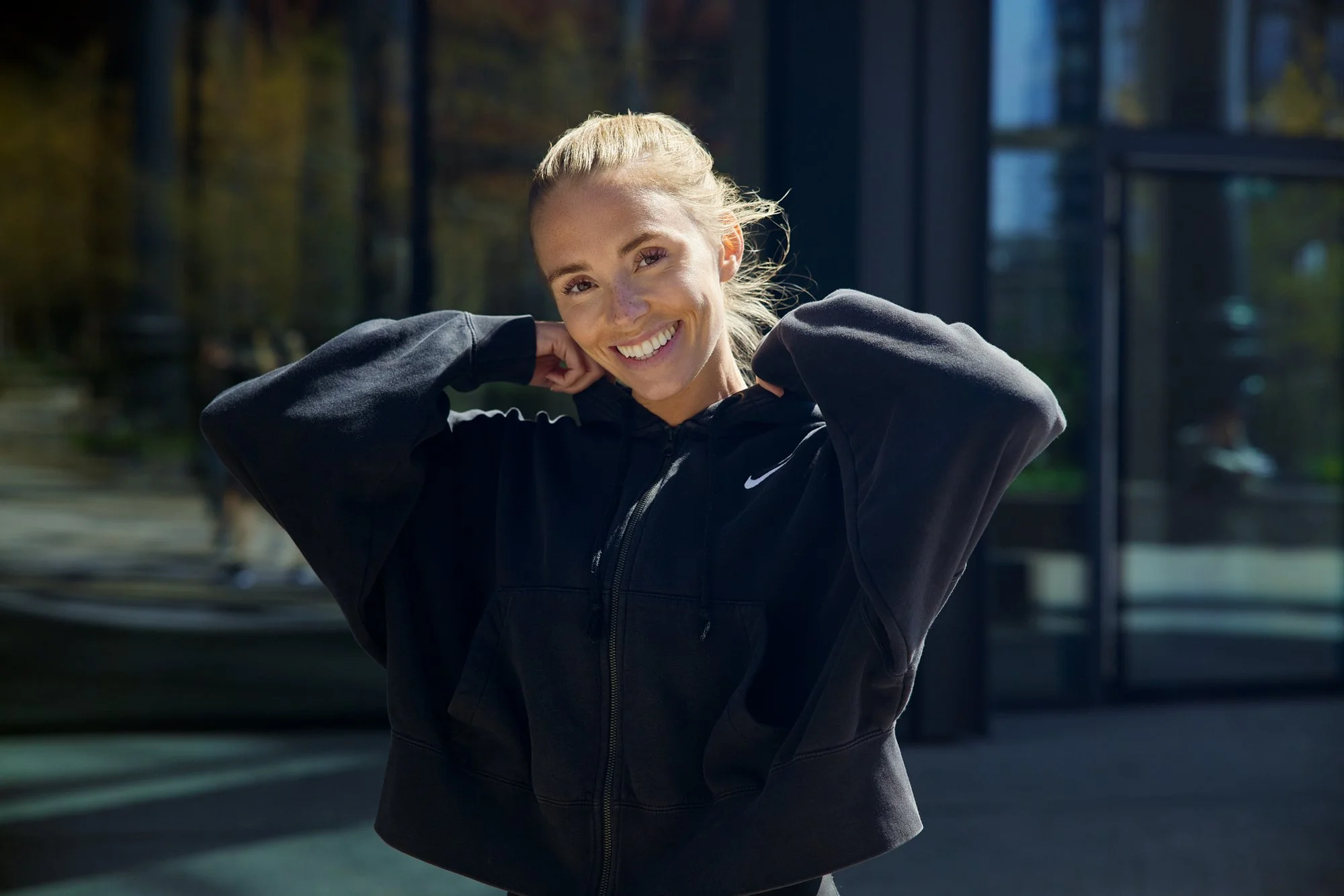Portrait of smiling girl wearing a black nike sports top and pulling up the collar with her hands. The sun lightens the right side of her face and her blond hair
