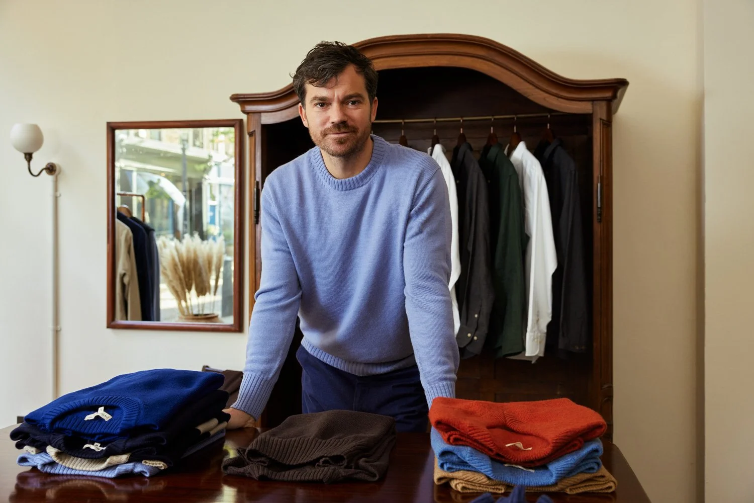 Callum of menswear brand, Original Fibres standing behind a table displaying neatly folded sweaters, with a clothing rack and a mirror in the background.