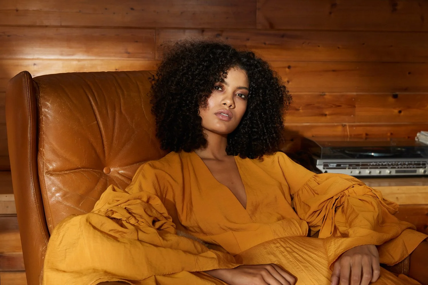 girl sitting on a vintage leather chair wearing an ochre cotton dress in the sunlight in a midcentury modern home