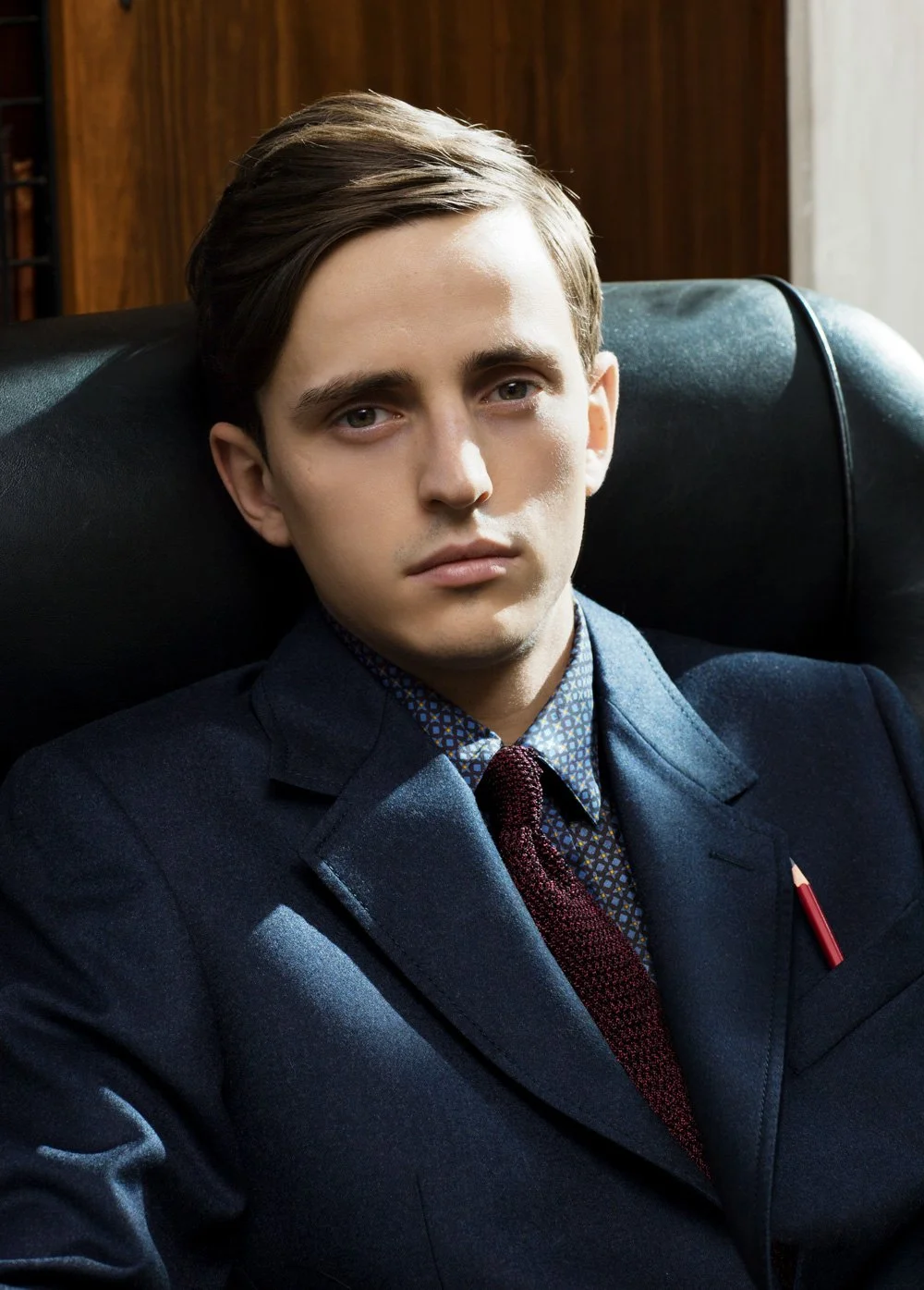male model sitting in a leather chair wearing a blue jacket, patterned shirt and maroon tie with a pencil in his breast pocket