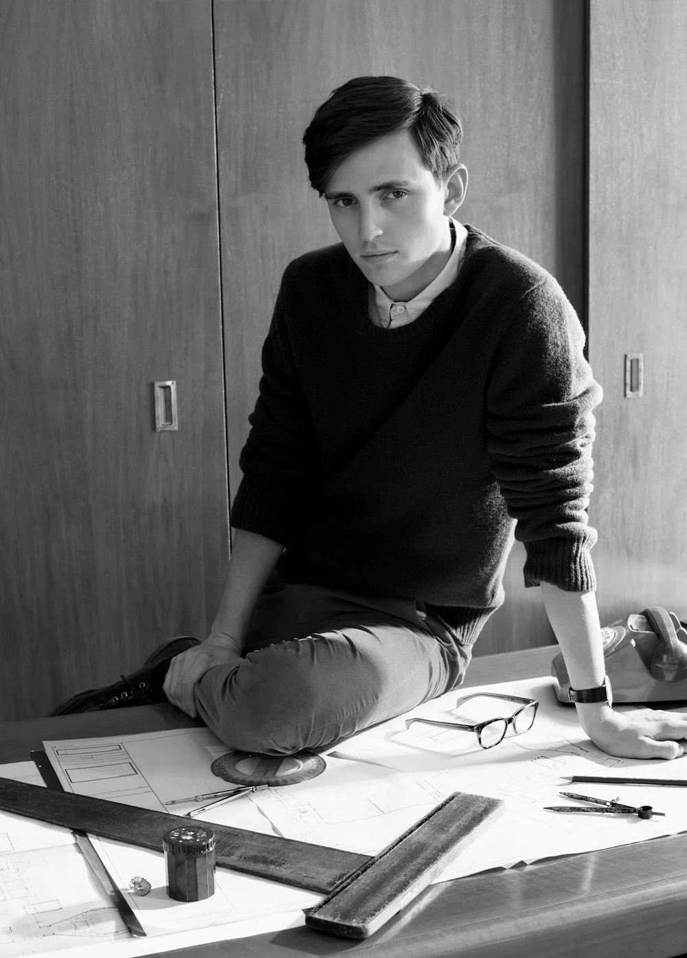 fashion image of man wearing a sweater shirt and trousers sitting on a desk with architects plans and a vintage dial telephone landline