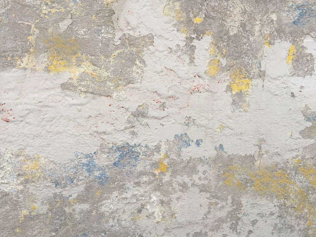 A close-up of a weathered wall with peeling paint in various colors including white, yellow, blue, and gray.
