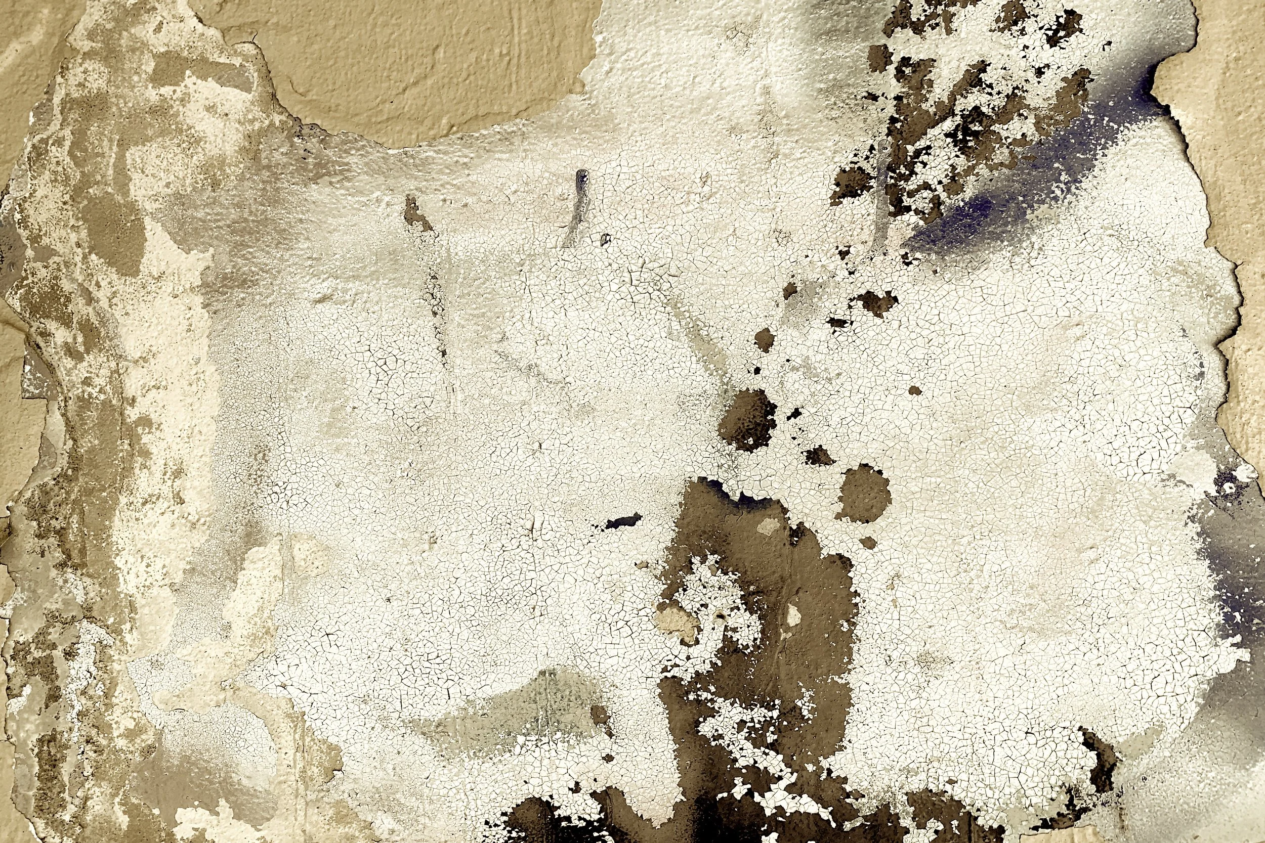 Close-up of peeling and cracked paint on a wall with beige, white, and dark brown patches.
