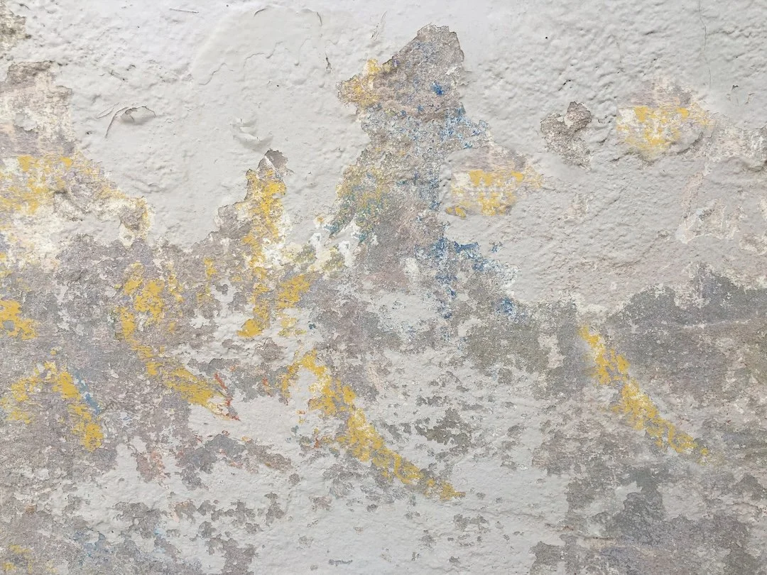 Close-up of a worn, peeling painted wall with patches of yellow, blue, and gray paint exposed.