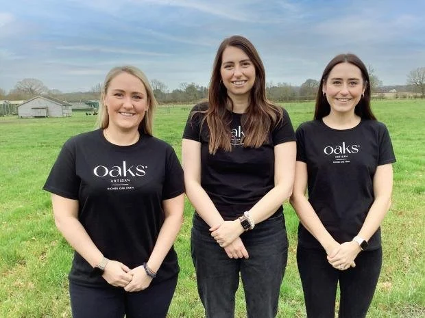 Three women standing outdoors on a grassy field, smiling, wearing black Oak's Artisan t-shirts, with a cloudy sky and rural landscape in the background.