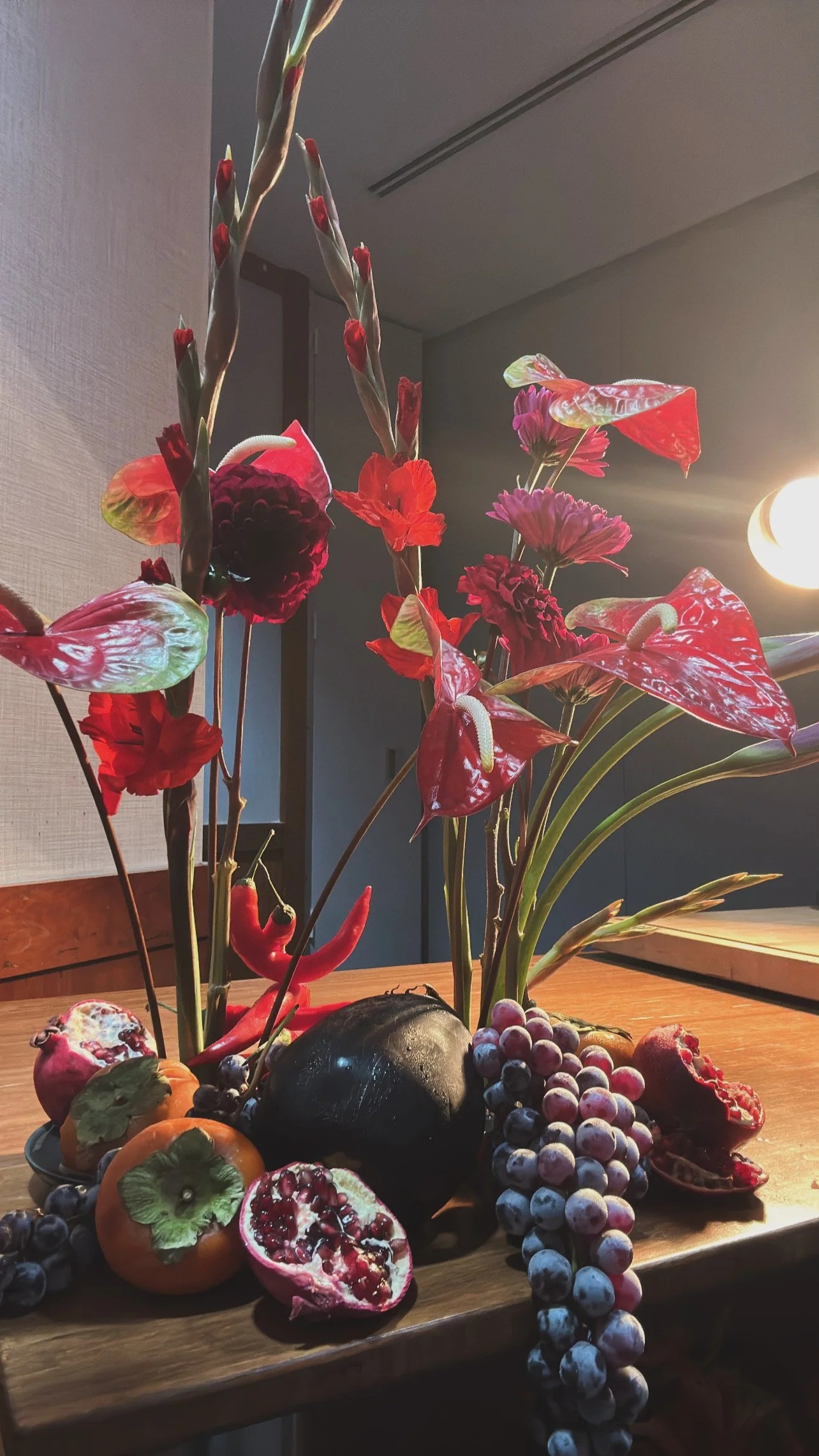 A floral arrangement with red and pink flowers, surrounded by an assortment of fruits including grapes, pomegranate, persimmon, and watermelon, placed on a wooden table.