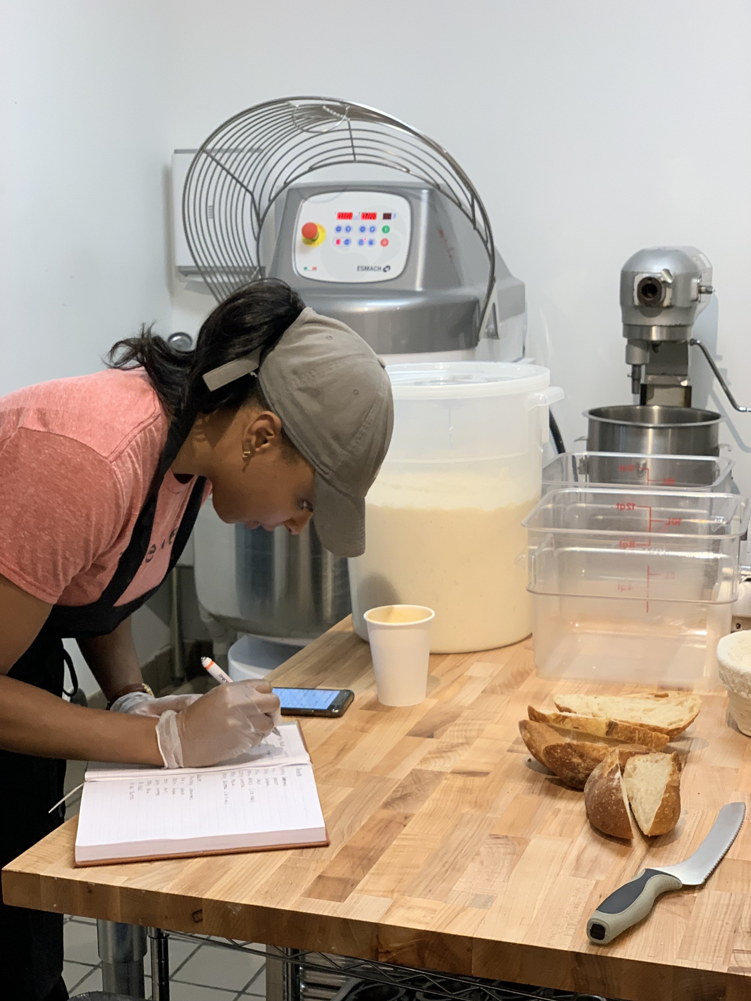 A person wearing a gray cap, gloves, and a pink shirt is writing in a notebook on a wooden counter. On the counter, there are slices of bread, clear plastic measuring containers, a large mixing bowl, and a white cup. In the background, industrial kitchen equipment including a large dough mixer and a dough proofer with digital displays are visible.