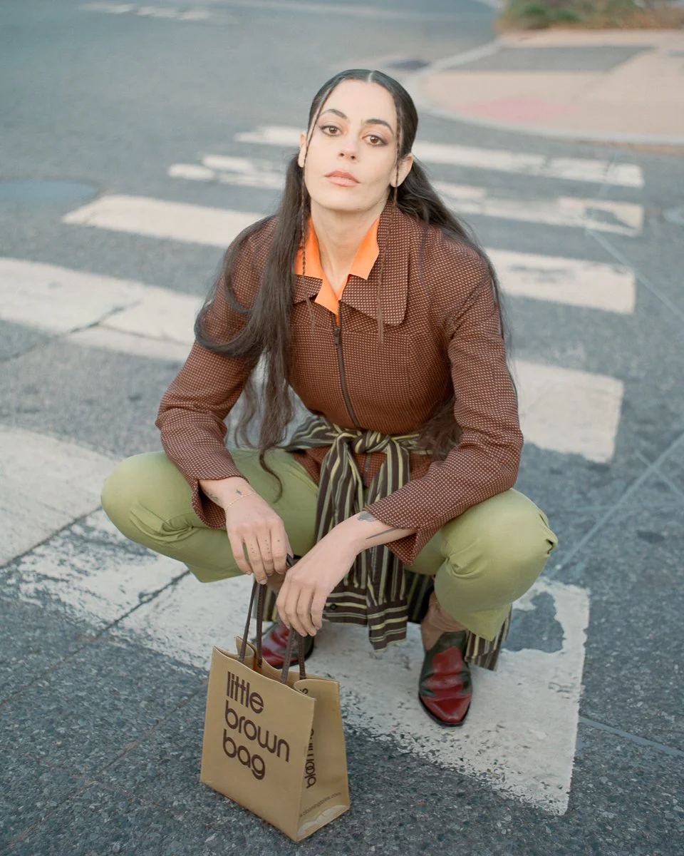 A woman is squatting on a crosswalk at an intersection. She has long dark hair, wears a brown jacket with an orange shirt underneath, light green pants, and red shoes. She is holding a beige shopping bag that says 'little brown bag'.