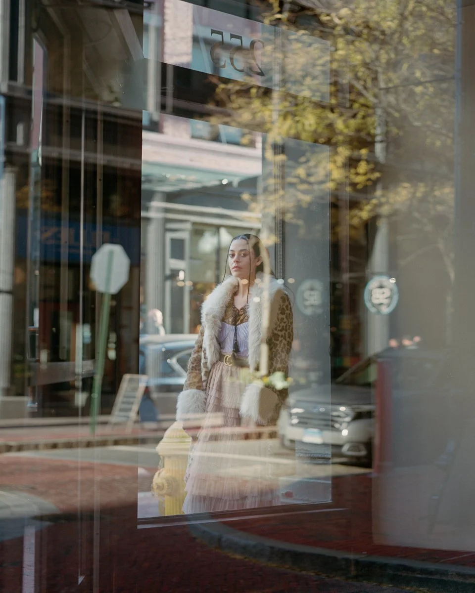 A woman standing outside looking through a glass window reflects the street scene with parked cars and trees with fall foliage.