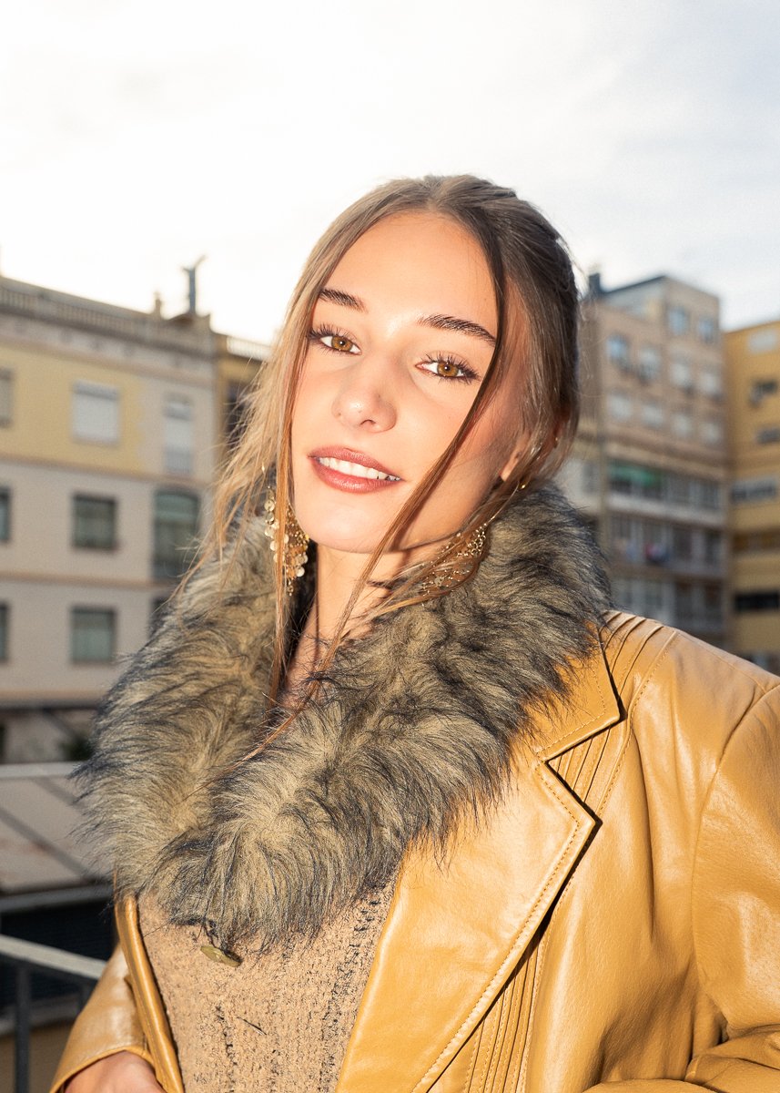 Young woman with light brown hair, hazel eyes, and earrings, wearing a tan leather jacket with a faux fur collar, outdoors in an urban setting during daytime.
