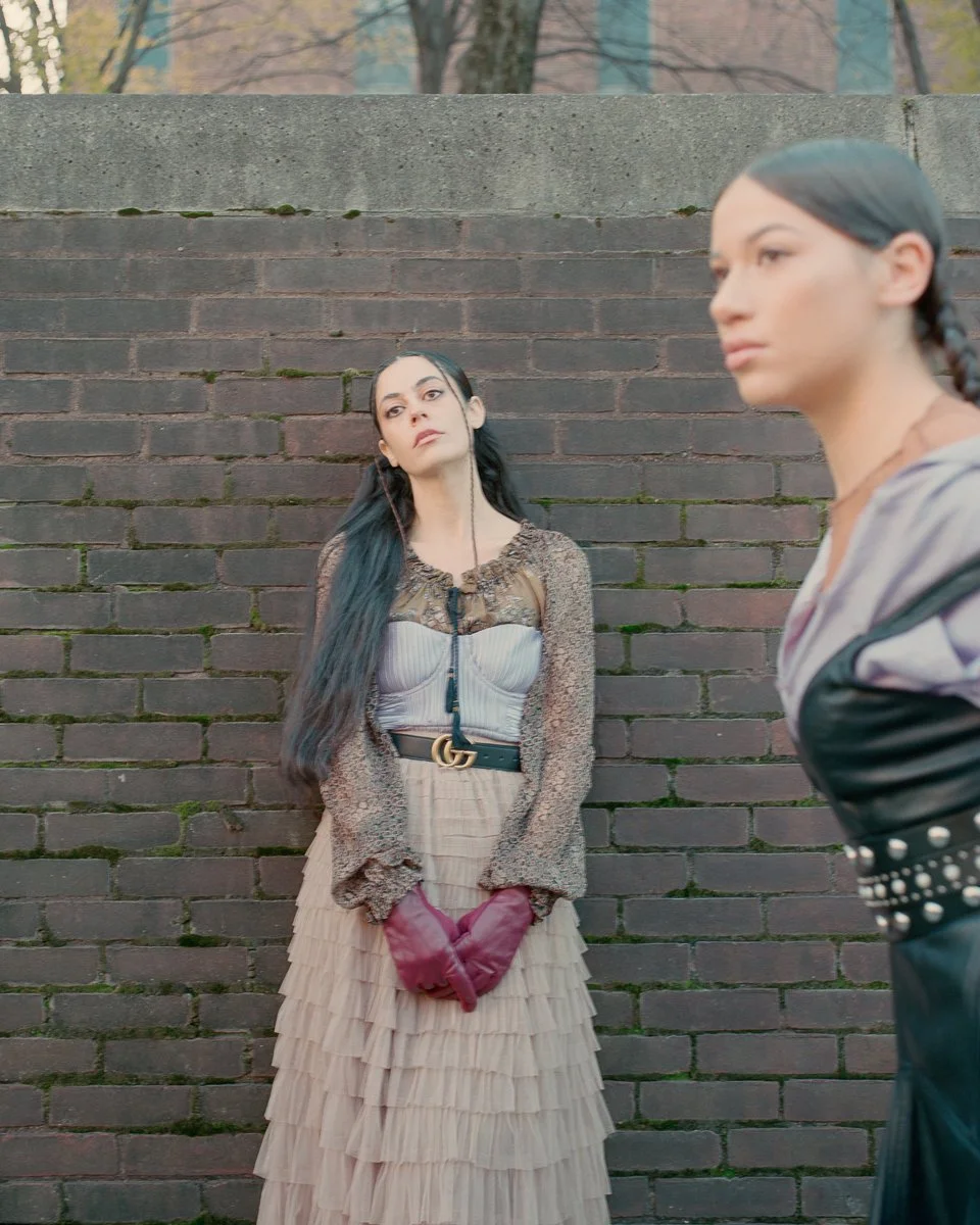 Two women stand against a brick wall outdoors. One woman in the foreground has blurred features and is looking to the right; she has dark hair in a braid, and is wearing a gray top with a black leather jacket with silver studs. The other woman in the