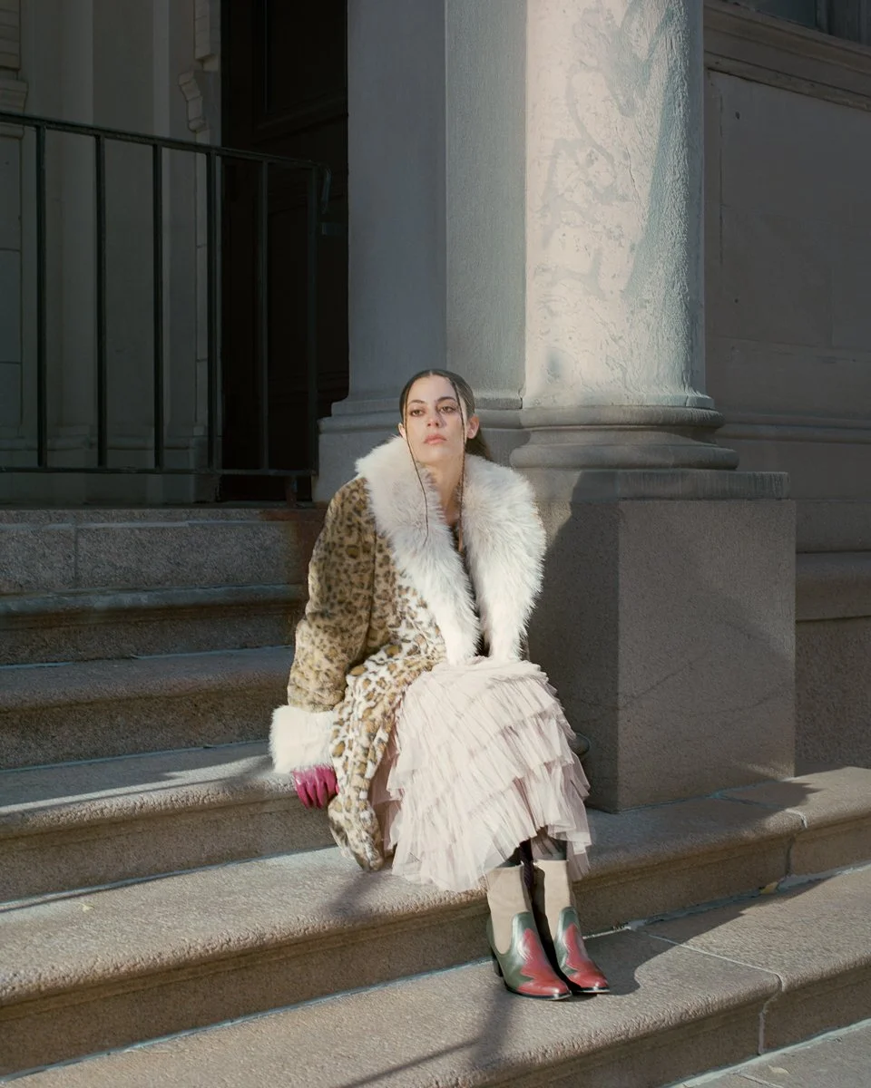 A woman sitting on steps outside a building, wearing a leopard print coat with white fur collar and cuffs, a flowy beige skirt, pink gloves, and cowboy boots.