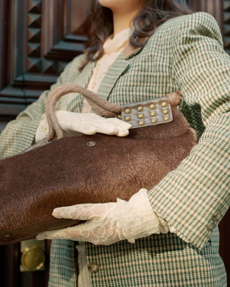A woman dressed in vintage clothing holding a brown leather purse with gloves, standing in front of an ornate wooden background.