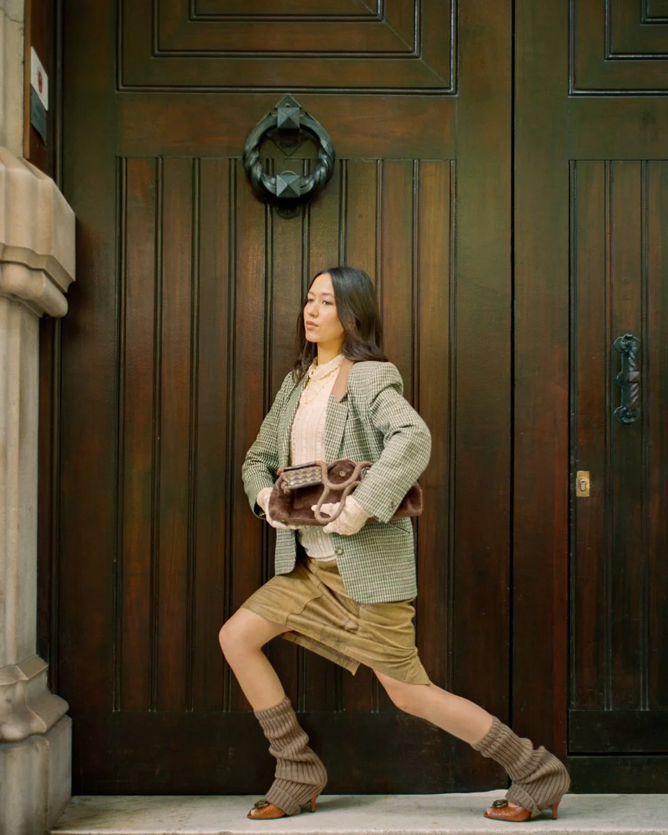 A woman in vintage-style clothing standing in front of a dark wooden door with her left knee bent and her right leg extended behind her, holding a brown purse.