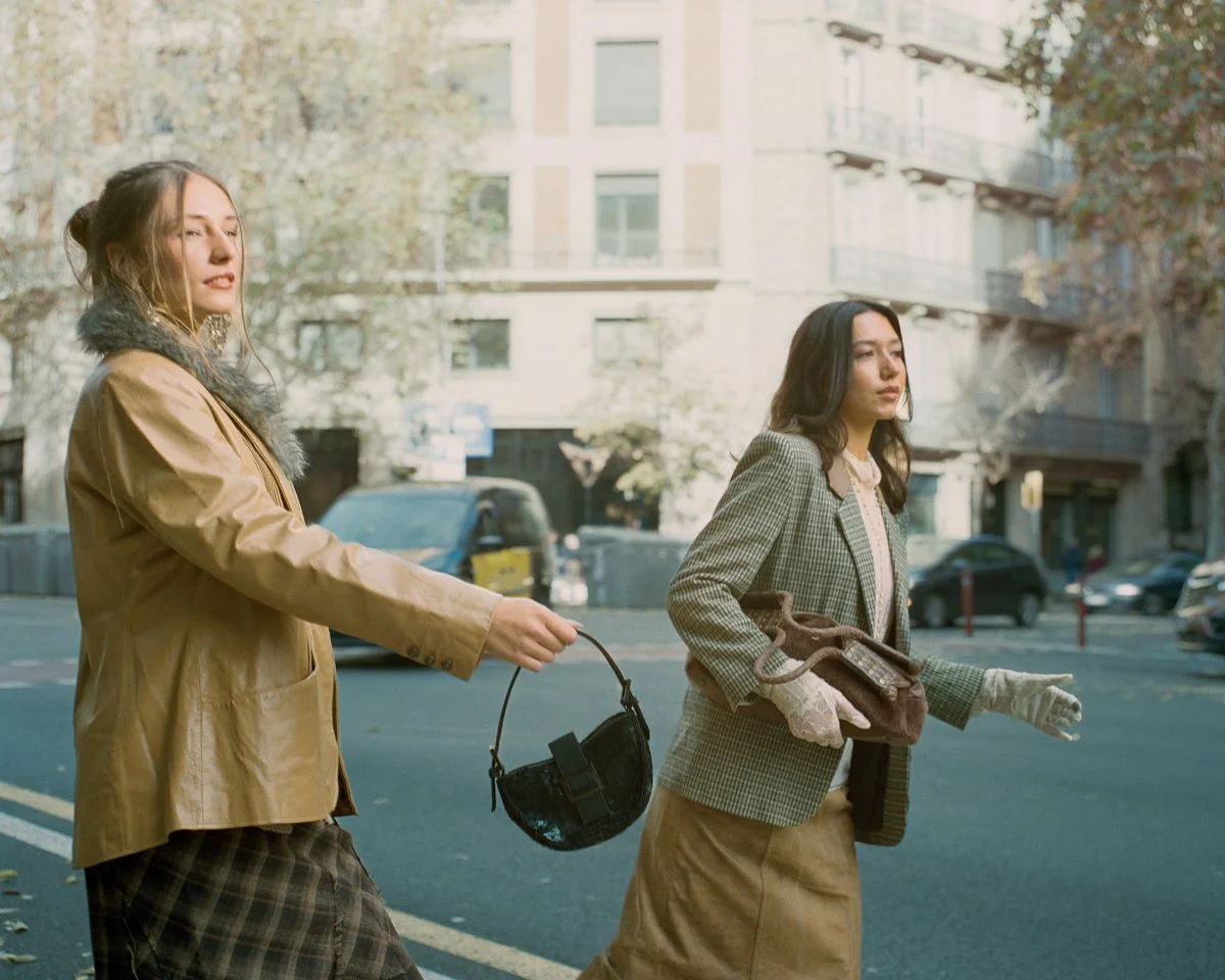 Two women walking on the street, with one holding a purse and the other holding gloves and a small bag, in an urban setting with buildings and cars in the background.