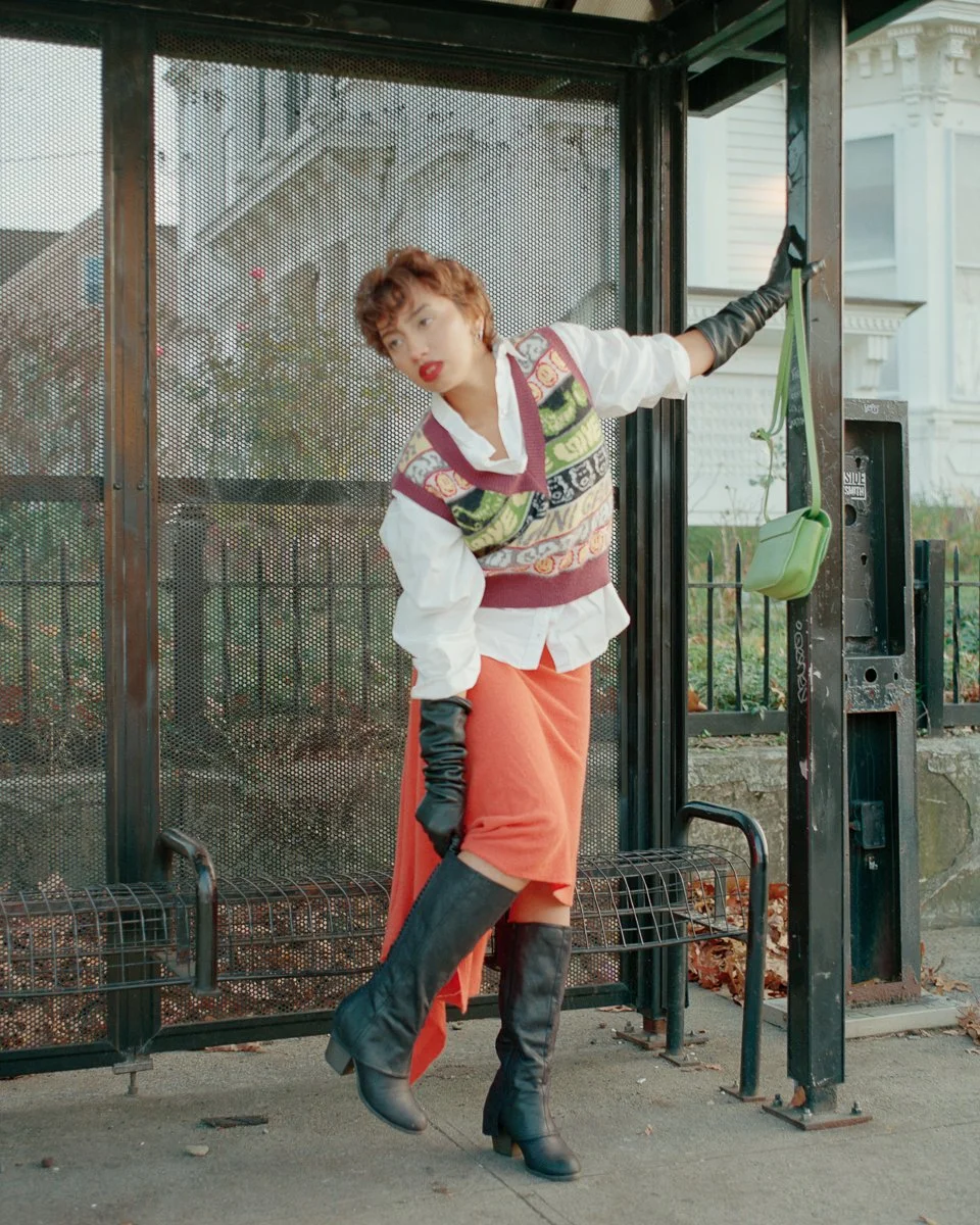 A woman with short curly hair, wearing a patterned sweater vest over a white shirt, orange skirt, black gloves, and tall black boots, standing at a bus stop and looking to the side.