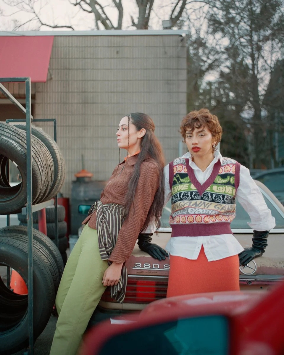 Two women leaning against a vintage car in an outdoor setting with tire racks nearby. One has long dark hair, wearing a brown top and light-green pants, while the other has curly short hair, wearing a colorful sweater vest over a white shirt and an o