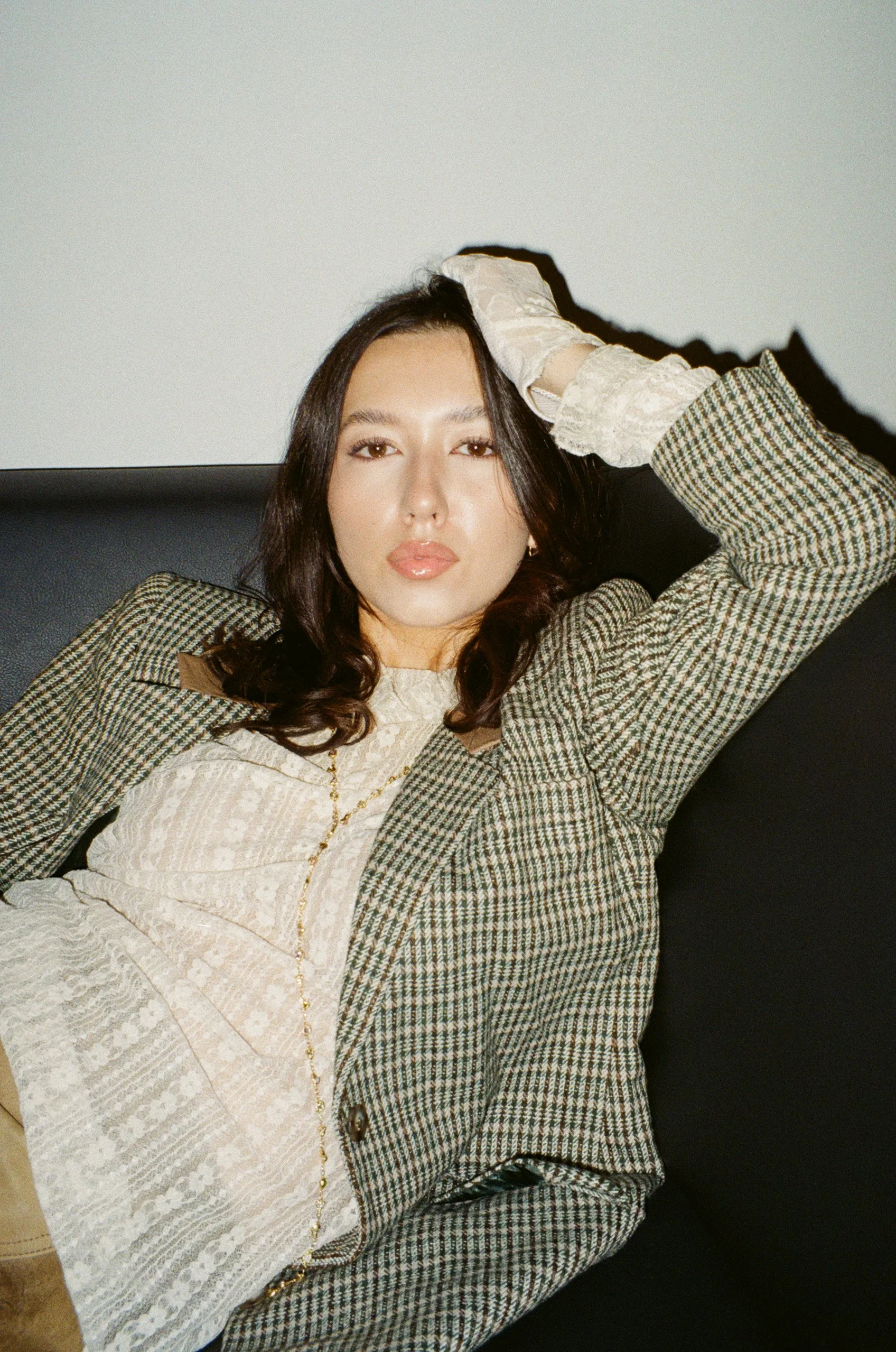 A woman with long dark hair, wearing a plaid blazer over a white lace top, reclining on a black sofa, looking directly at the camera with one hand resting on her head.