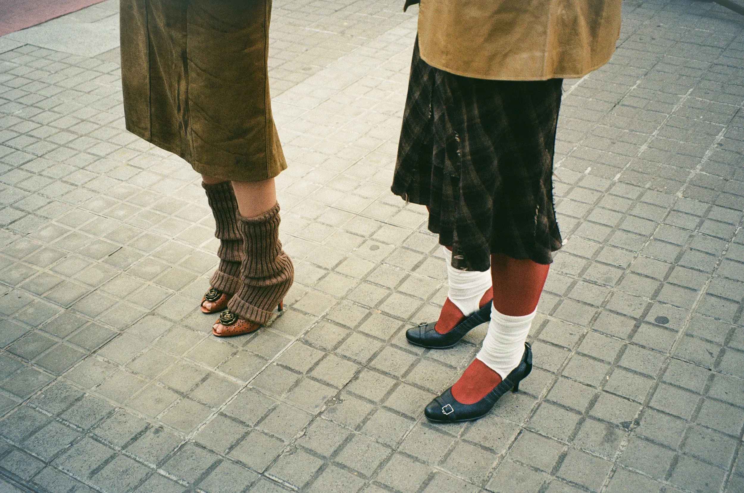 Close-up of two women standing on a tiled sidewalk, showing their legs and shoes.