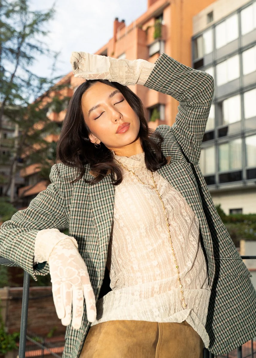 A woman with dark hair and light skin, wearing a checkered blazer and lace top, stands outdoors with her eyes closed, one hand on her forehead, in a city neighborhood with apartment buildings and trees in the background.
