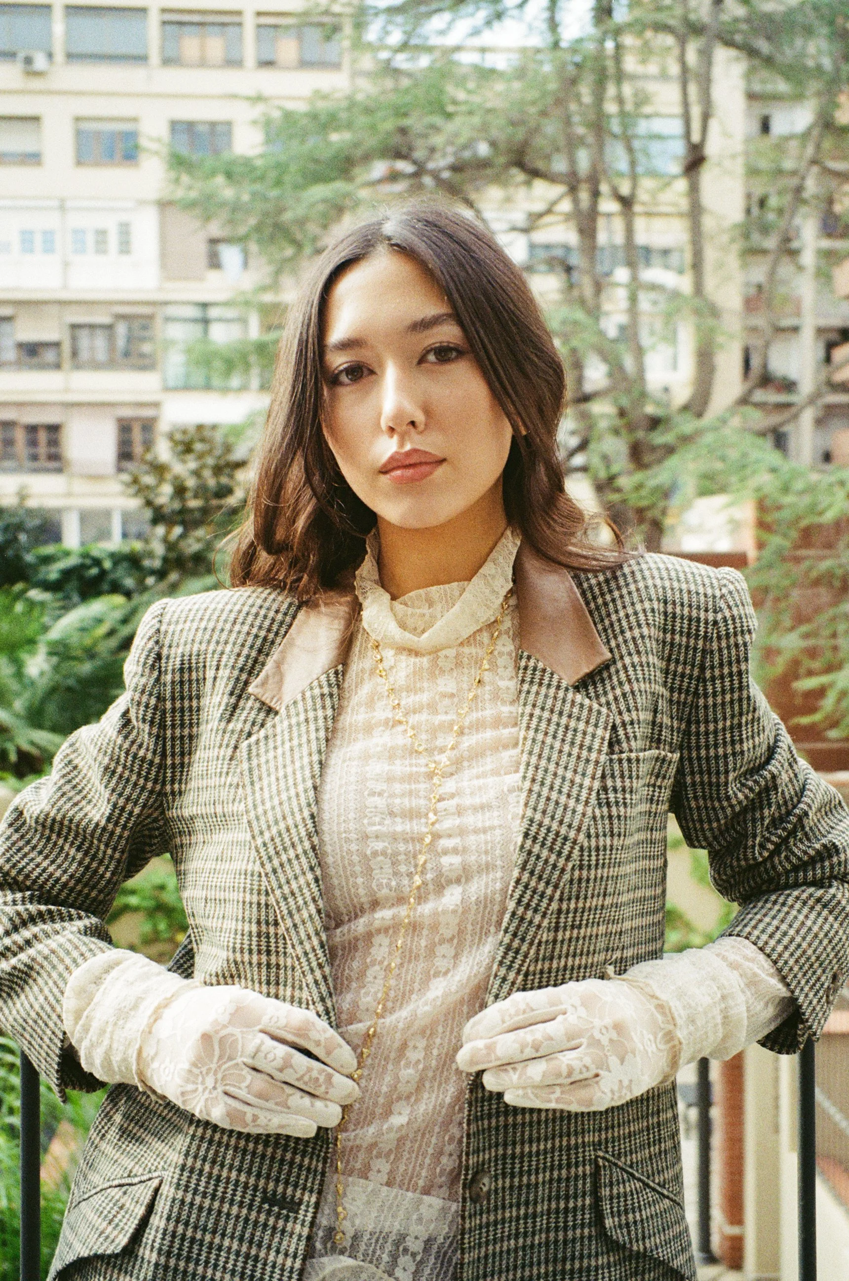 A young woman with shoulder-length dark hair, dressed in vintage clothing, stands outdoors on a balcony with greenery and apartment buildings in the background.