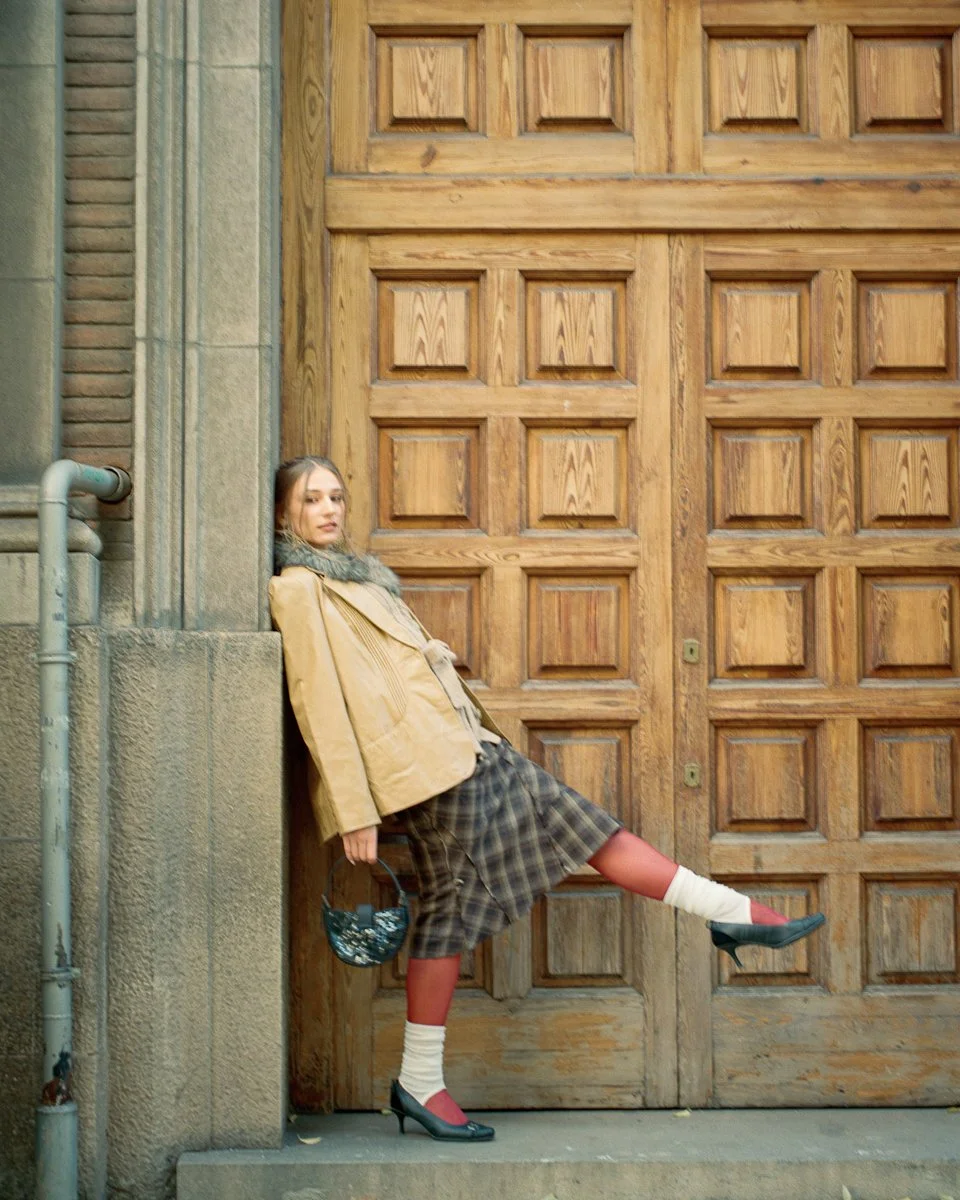 A woman standing against a wall with a large wooden door, wearing a beige jacket, plaid skirt, white socks, and high heels, holding a small handbag.