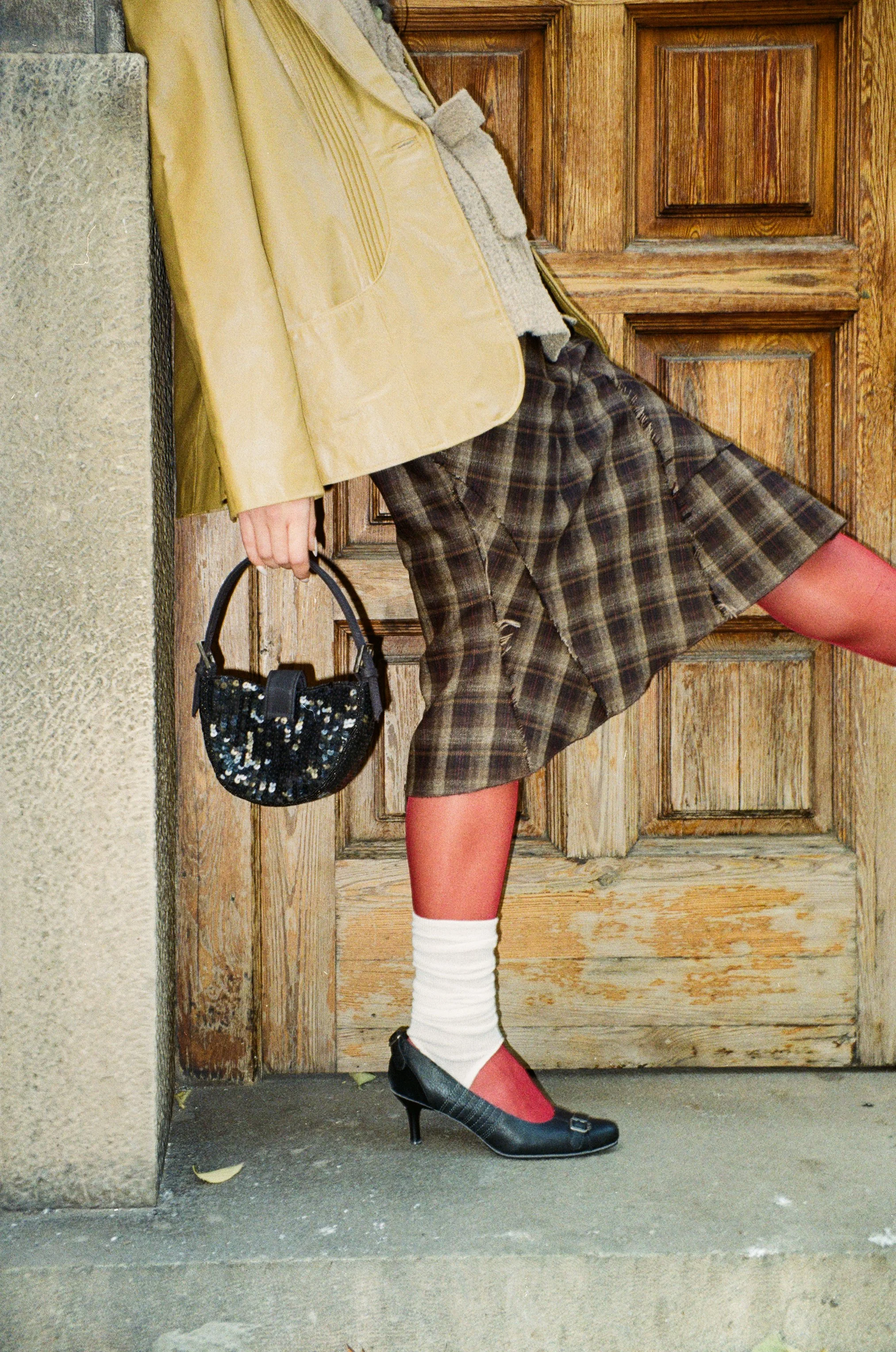 Close-up of a person's lower body wearing a plaid skirt, black high heels, white socks, red tights, and holding a small black sequined handbag, standing in front of a wooden door.
