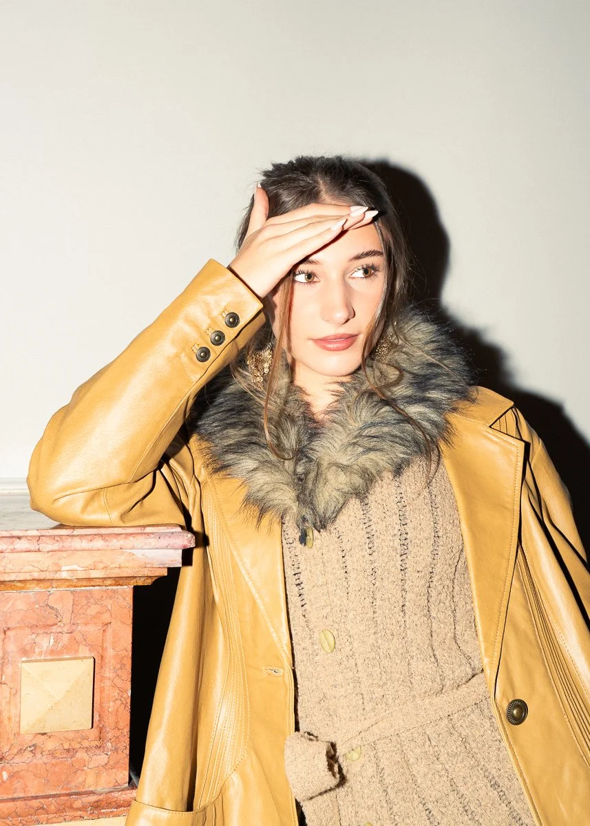 Young woman with dark hair, dressed in a tan leather coat with fur collar, leaning on a marble surface against a neutral background.