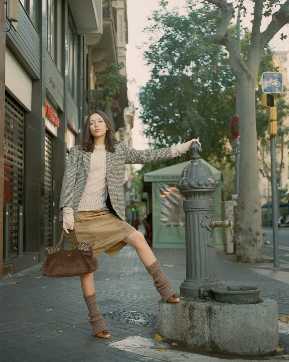 A woman in a plaid blazer, beige sweater, brown skirt, and knee-high socks stands on a city sidewalk, holding onto a vintage water fountain.