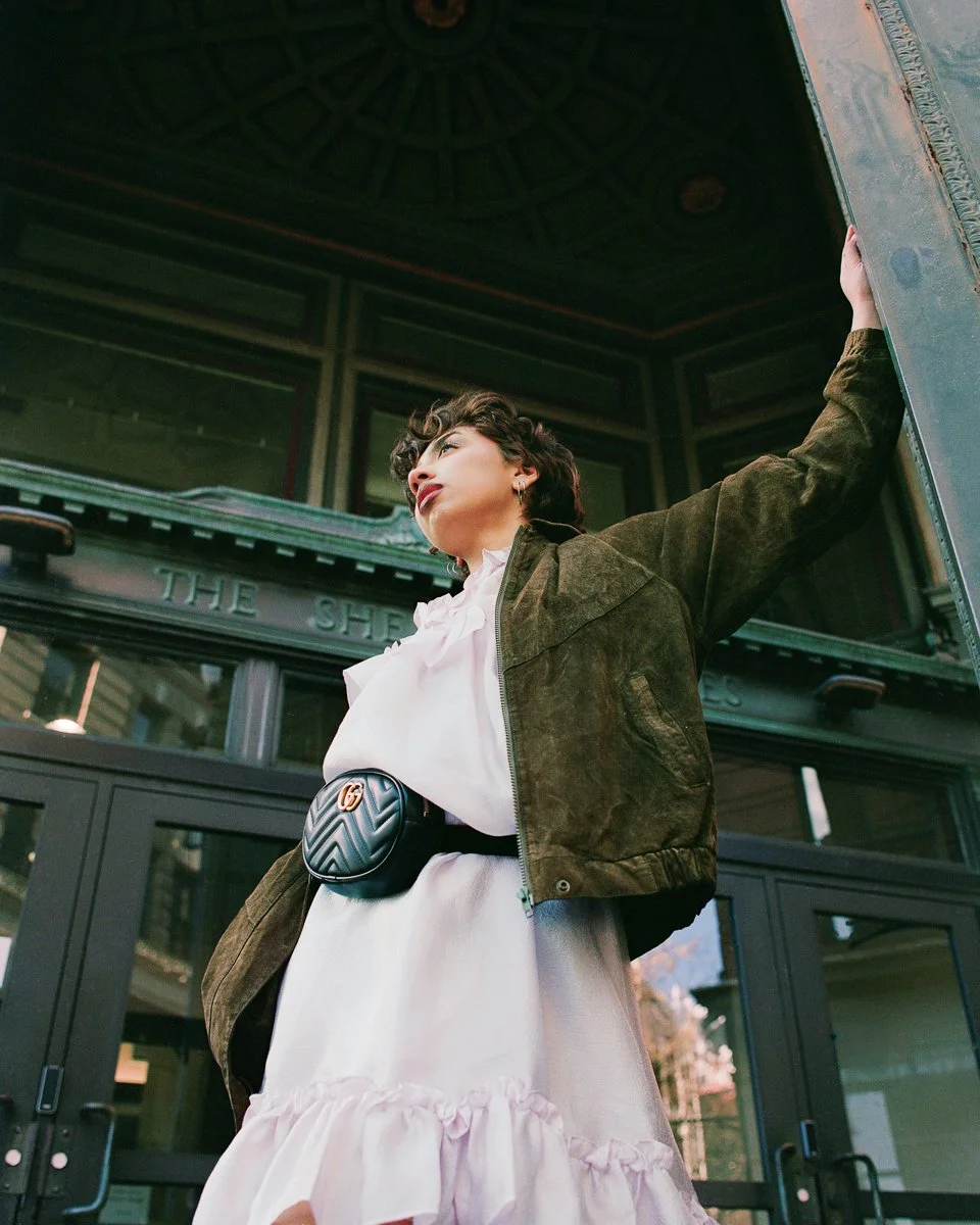 Woman wearing a white dress, olive green jacket, and a small black Gucci belt bag standing outside a building with glass doors.
