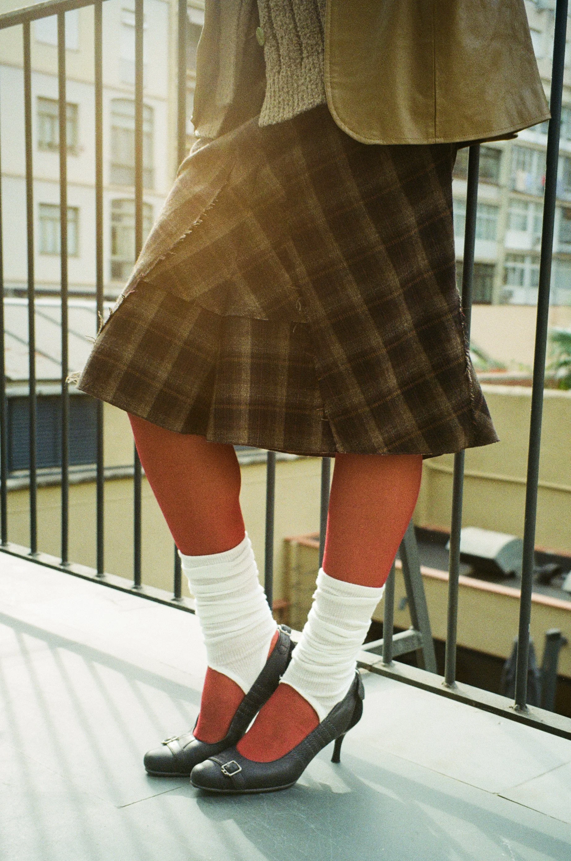 Close-up of a person standing on a balcony, wearing a plaid skirt, red tights, white leg warmers, black heels, and a brown jacket with an urban city background.