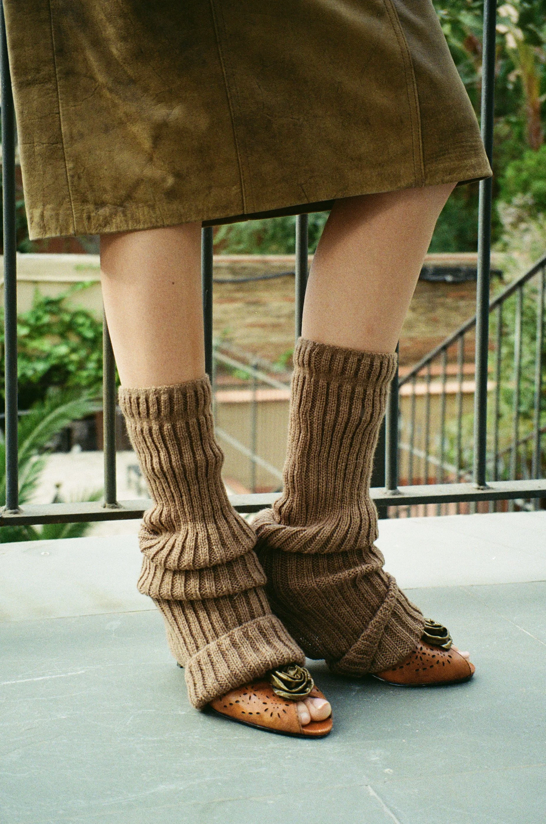 Close-up of a person wearing beige knee-high knitted socks, a brown skirt, and open-toed brown shoes with floral embellishments, standing on a balcony with a green outdoor background.