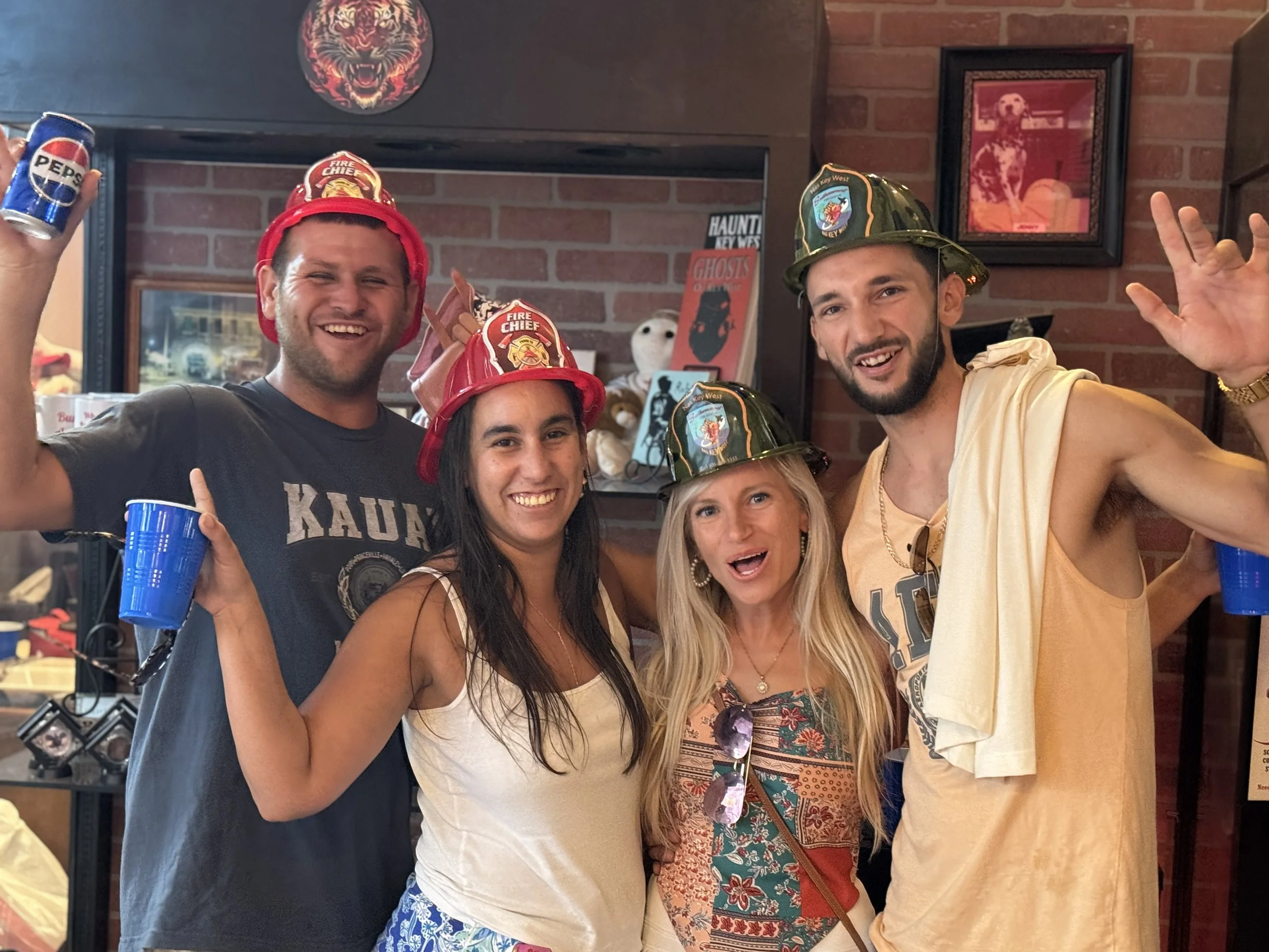 Group of five friends wearing firefighter hats, smiling and raising cans of Pepsi, at a party in a brick-walled room.