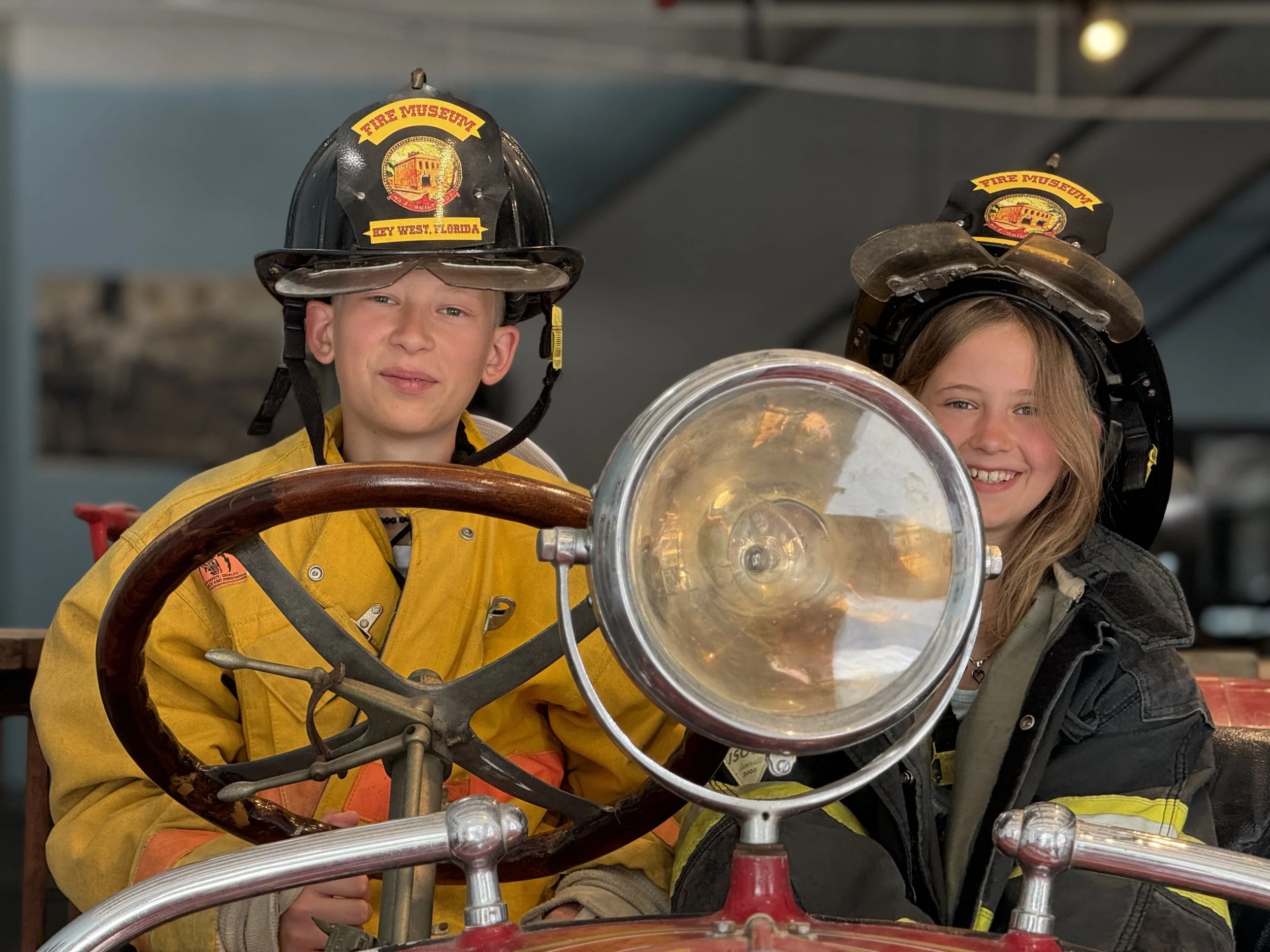 Two children dressed as firefighters wearing helmets, sitting on a fire truck with a large silver headlamp and a vintage steering wheel, smiling at the camera.
