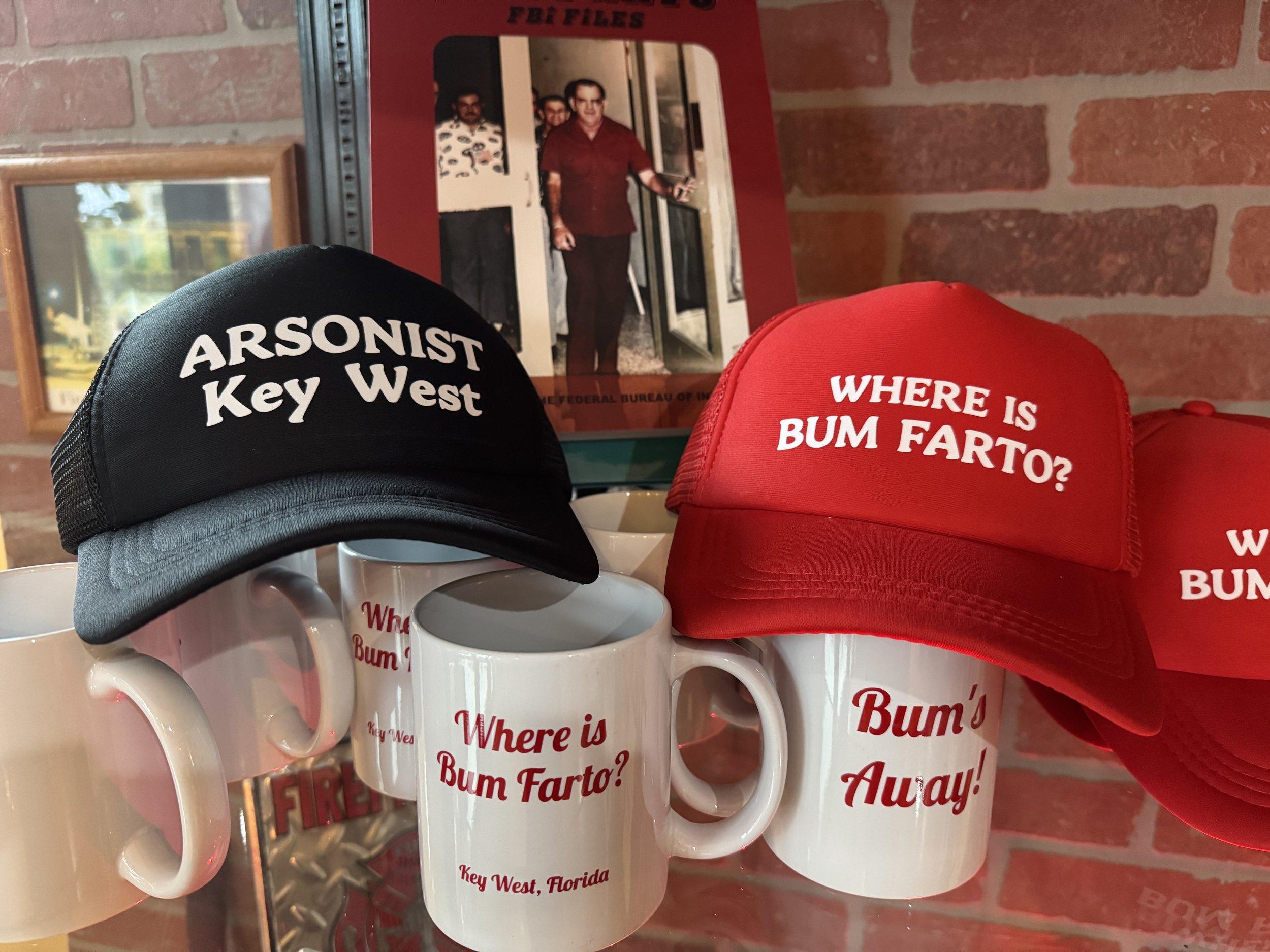 Black and red baseball caps with humorous text, placed on top of white coffee mugs with matching phrases, against a brick wall background with framed pictures.
