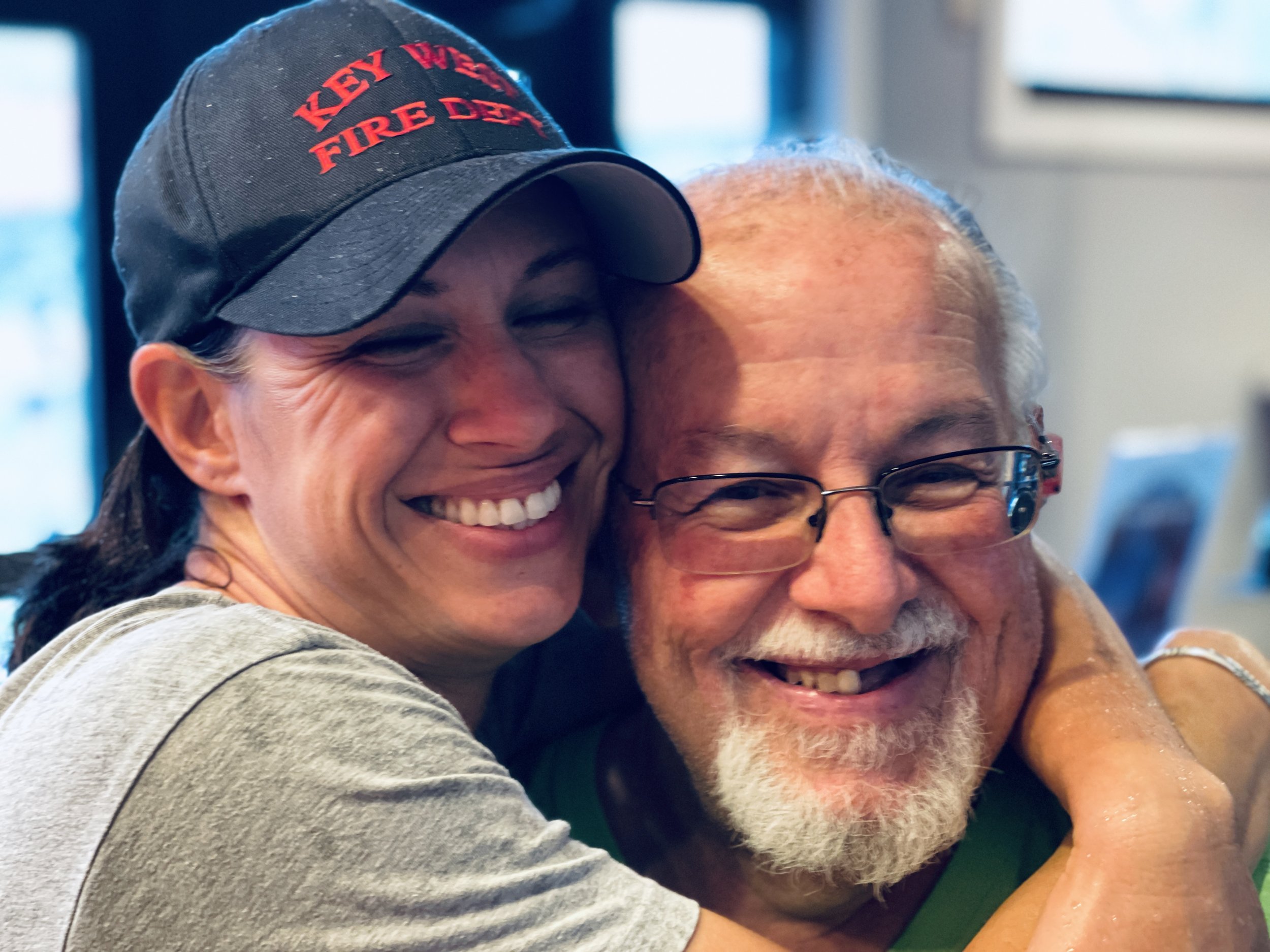 A woman wearing a black baseball cap with red text and a gray shirt is hugging a man with glasses and a gray beard. Both are smiling and appear happy.