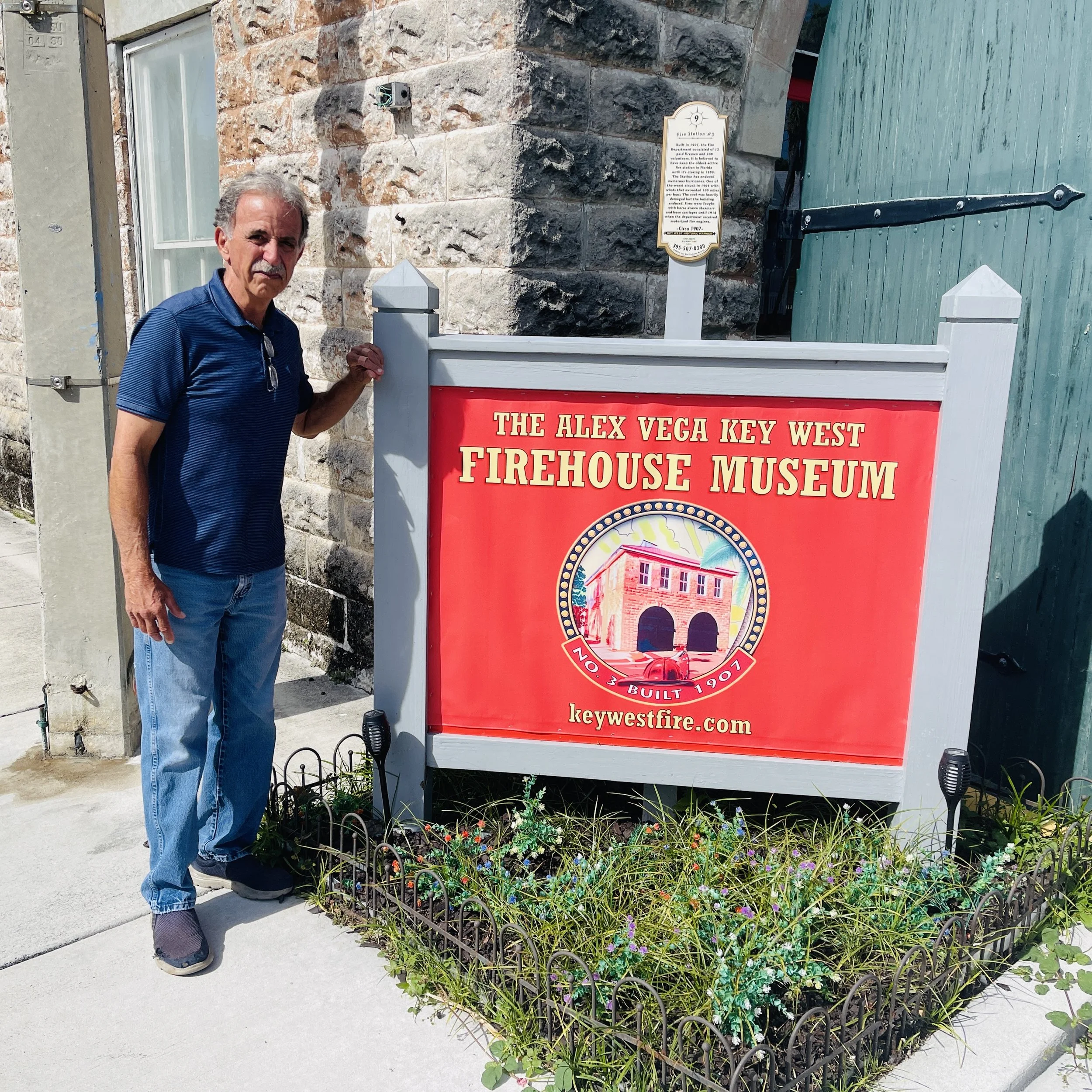 Captain Alex Vega standing outdoors next to a sign for the Alex Vega Key West Firehouse Museum, with a stone building and a wooden green door behind him.