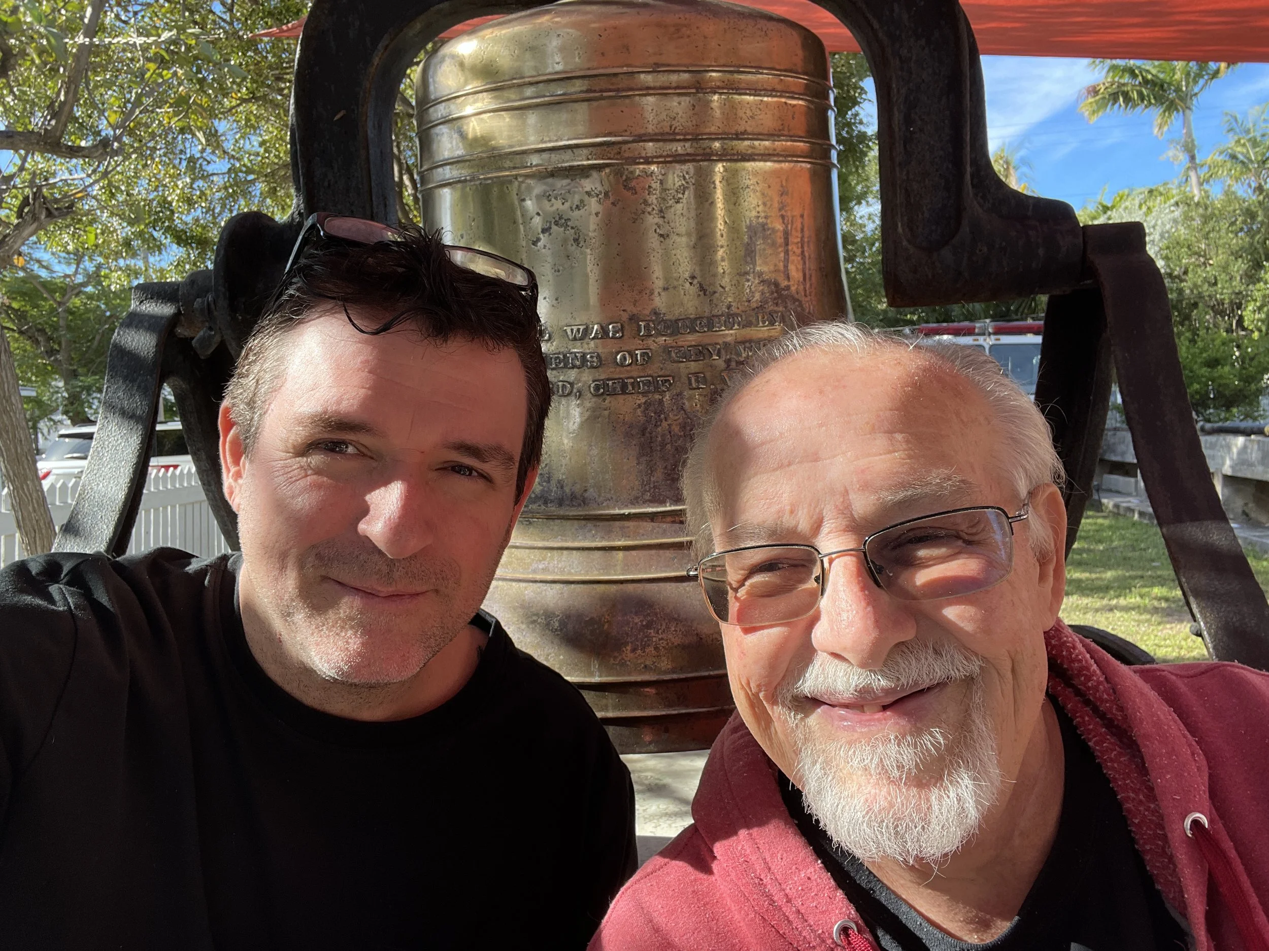 Two men taking a selfie outdoors, standing in front of a large, rustic, metal bell hanging from a structure. The younger man on the left has dark hair and is wearing a black shirt, the older man on the right has white hair, a beard, and glasses, and is wearing a red jacket. The background shows trees and a blue sky.