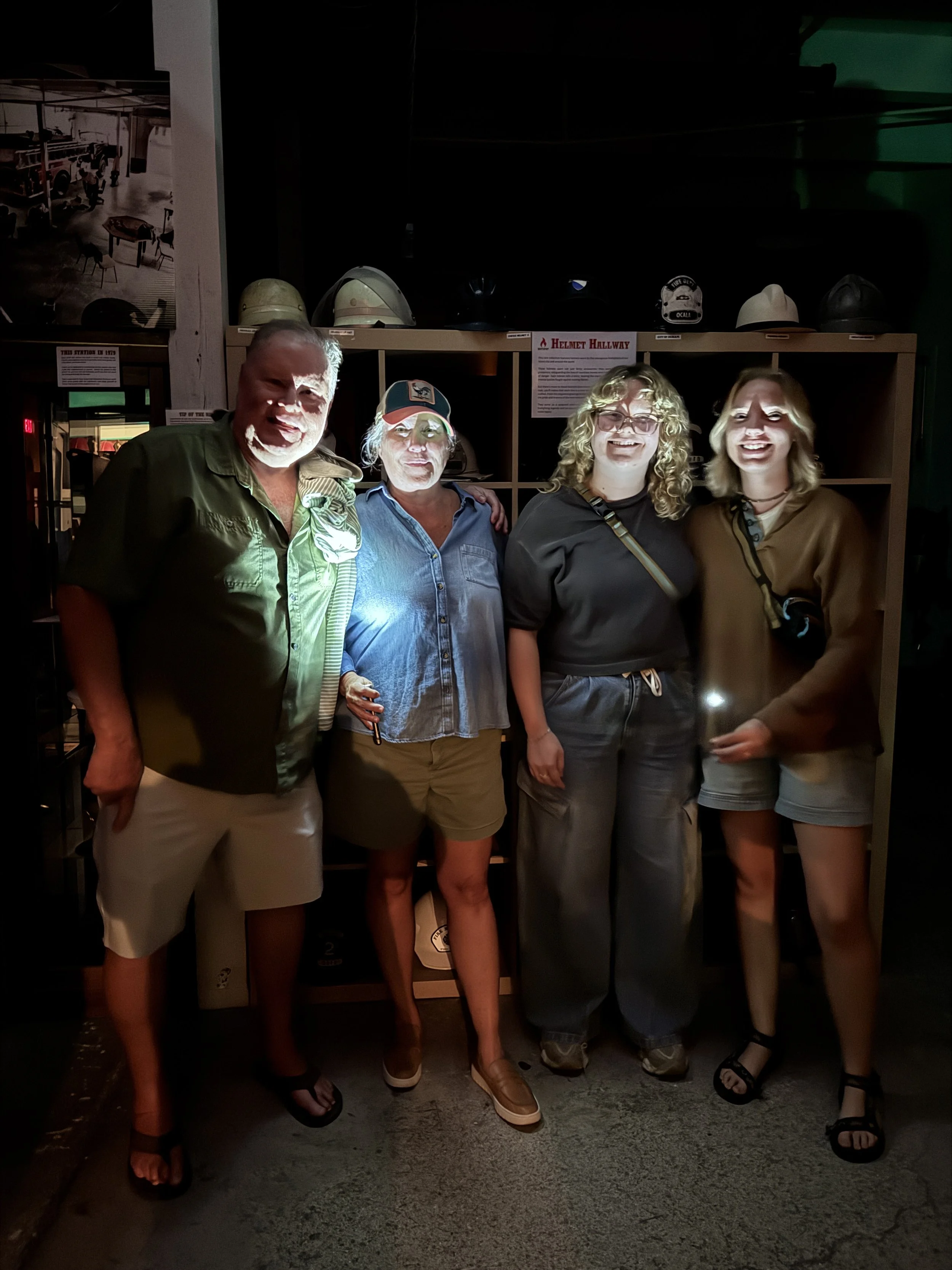 Four people standing indoors at night, posing for a photo with helmets displayed on shelves behind them.