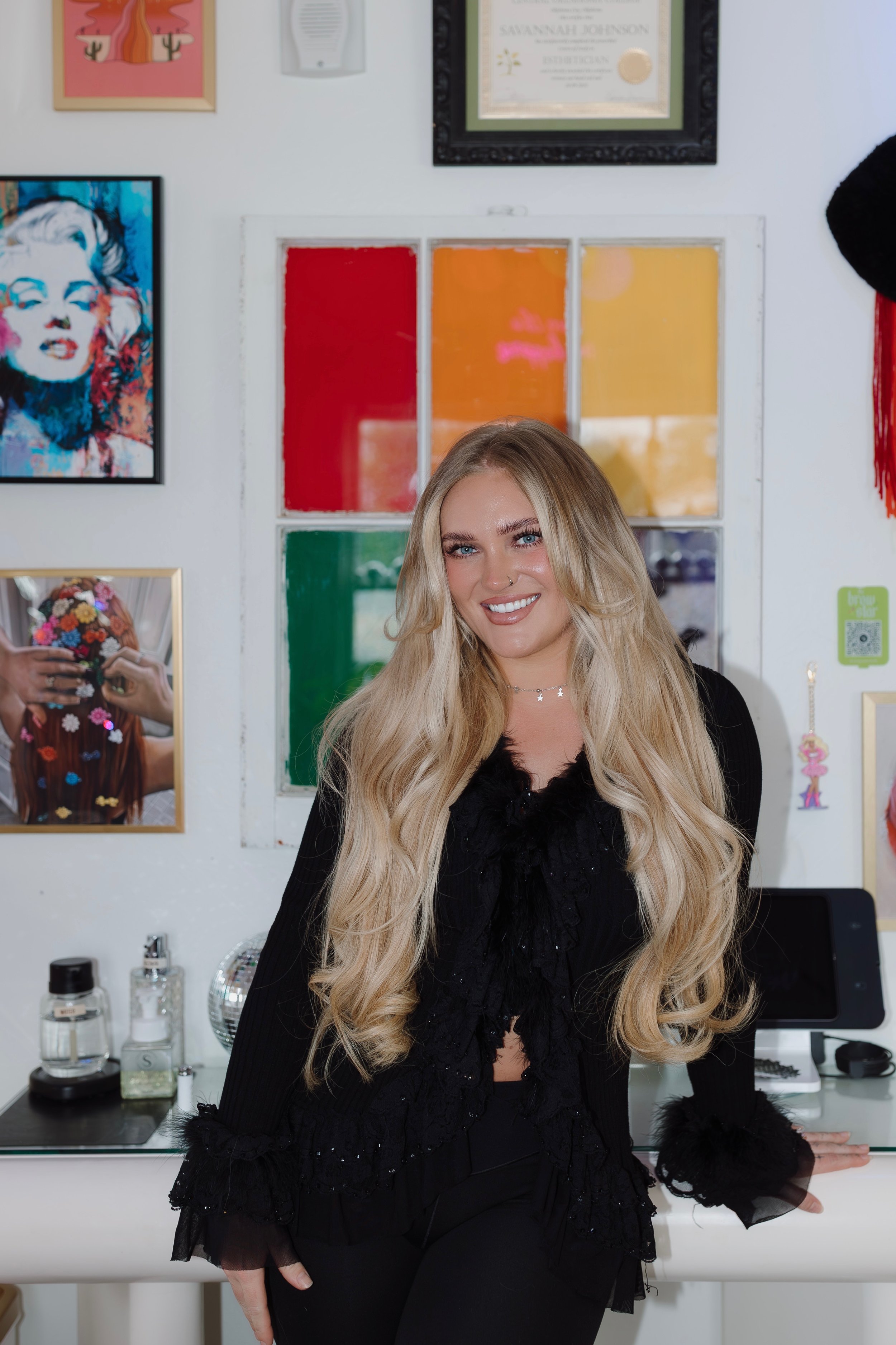 A woman with long blonde hair and blue eyes smiling, wearing a black outfit with fur cuffs, standing in a room with colorful artwork and a glass window behind her. There are various bottles and items on a counter in front of her.