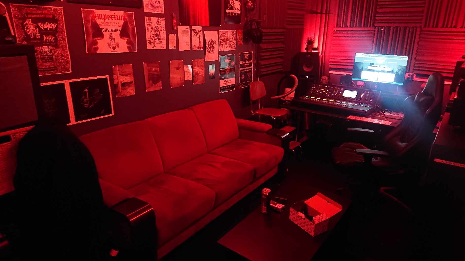 A music studio with red lighting, featuring acoustic foam panels on the walls, a desk with audio equipment and a computer monitor, a red sofa, and various posters.